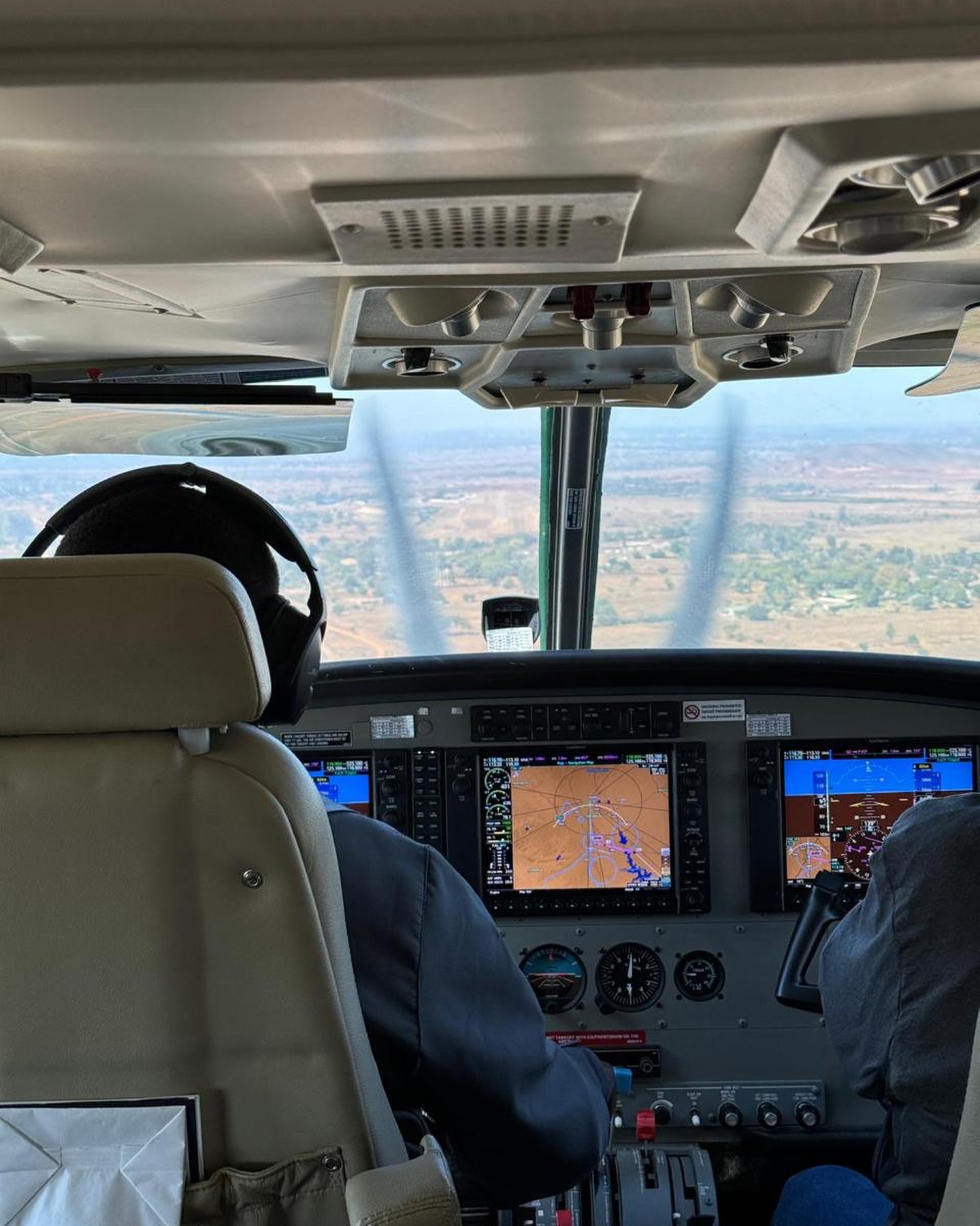 View from a small aircraft cockpit with pilots and glowing instrument screens, looking out to a wide landscape.