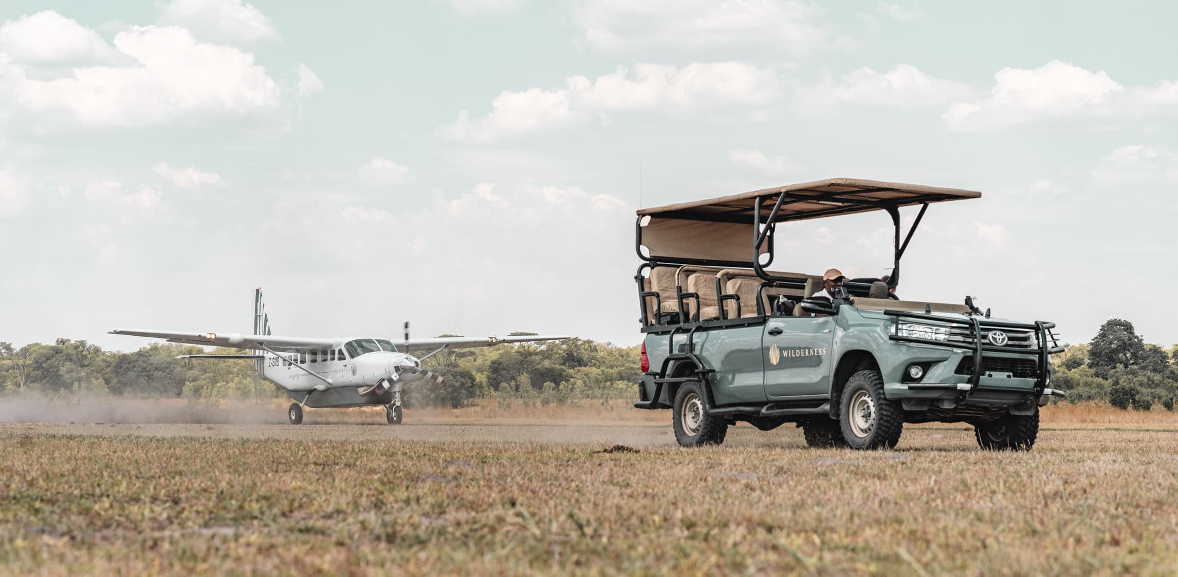 An open safari vehicle waits beside a dirt airstrip as a small plane taxis in the distance under pale clouds.