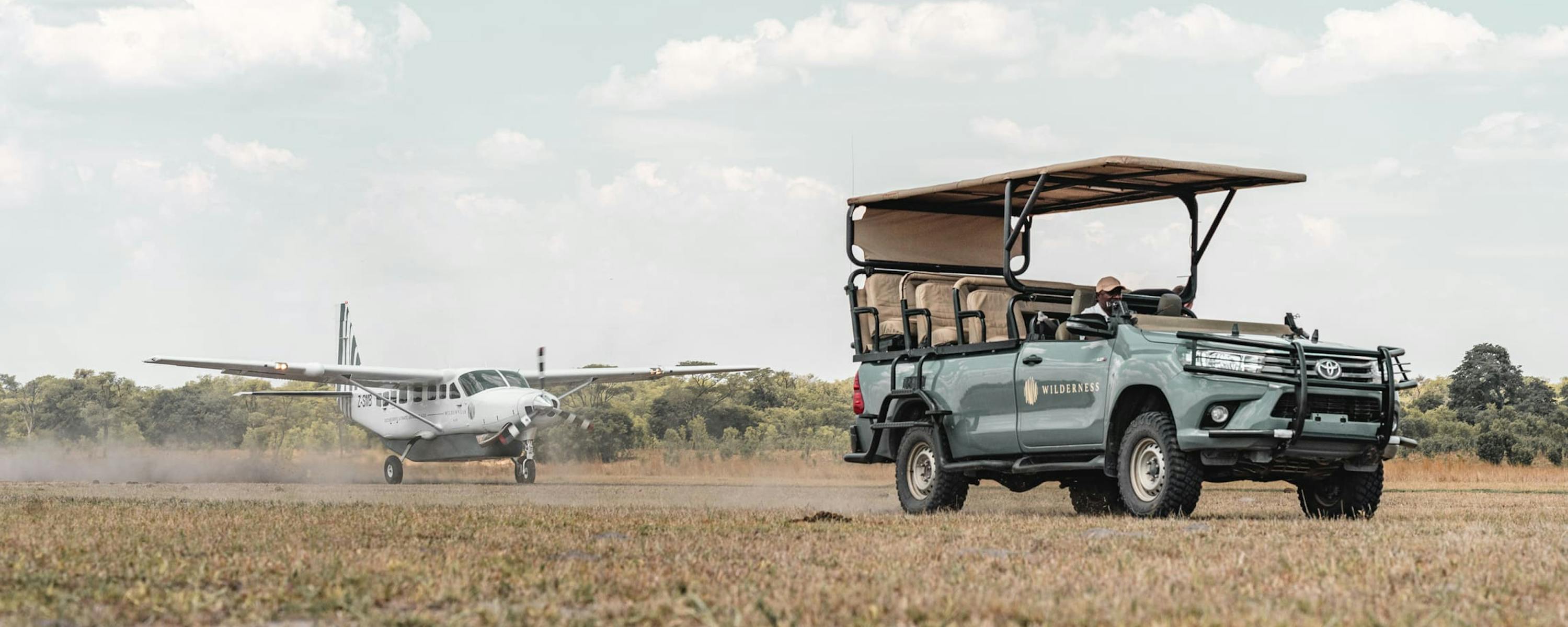 An open safari vehicle waits beside a dirt airstrip as a small plane taxis in the distance under pale clouds.
