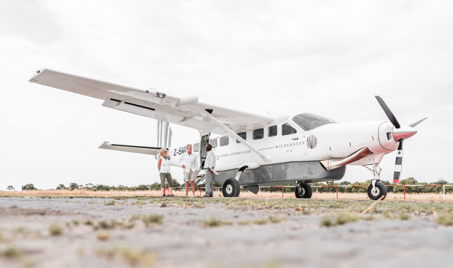 A small propeller plane is parked on a gravel airstrip, its white fuselage set against a wide gray sky overhead.