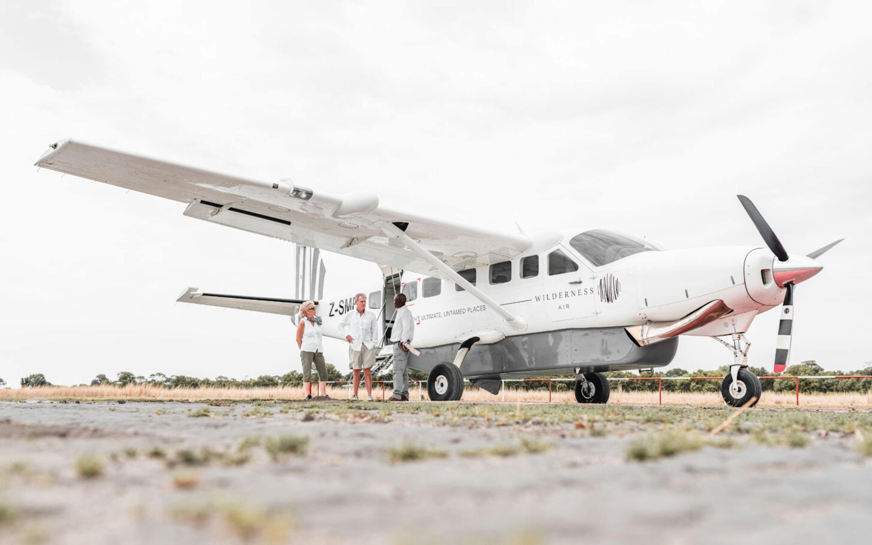 A small propeller plane is parked on a gravel airstrip, its white fuselage set against a wide gray sky overhead.