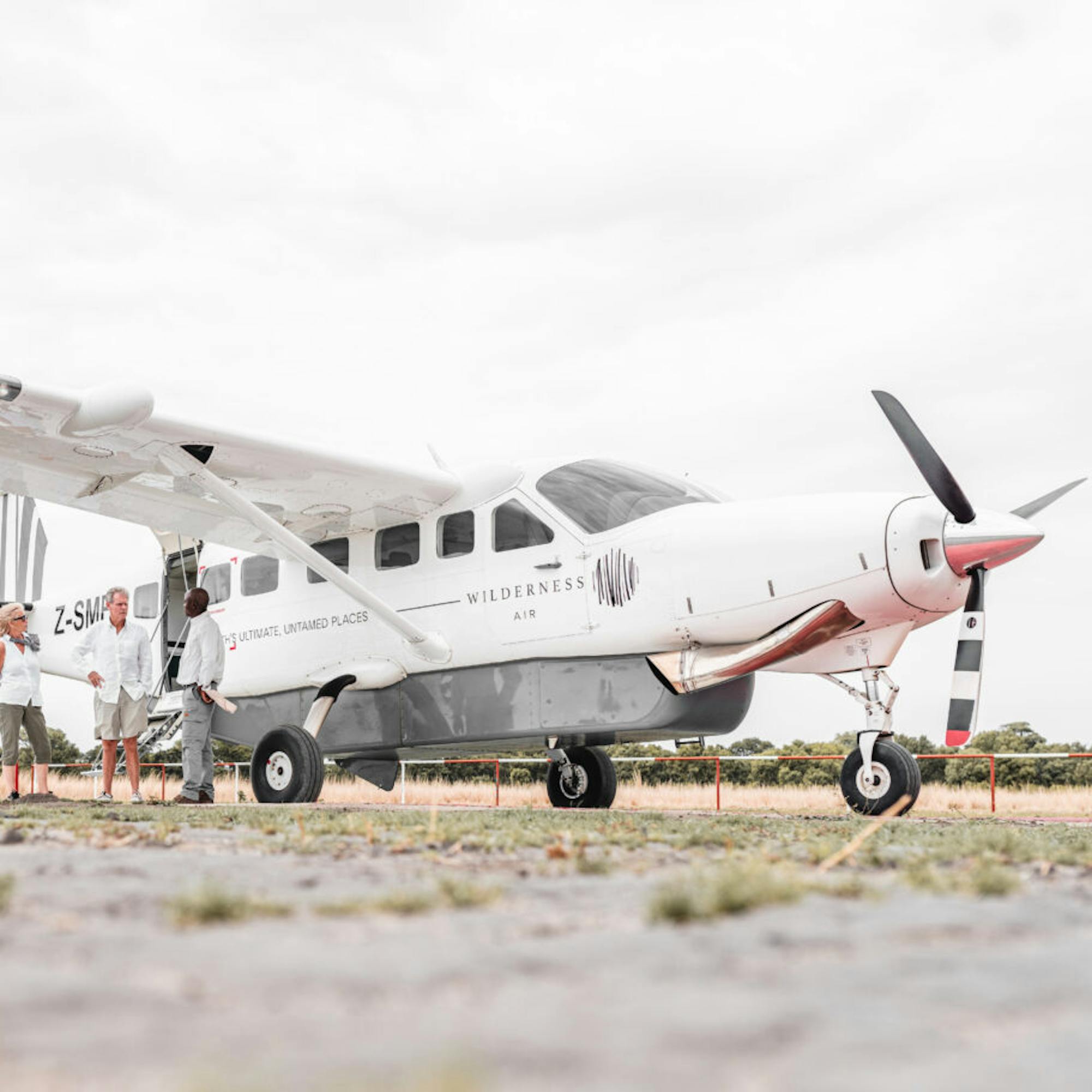 A small propeller plane is parked on a gravel airstrip, its white fuselage set against a wide gray sky overhead.