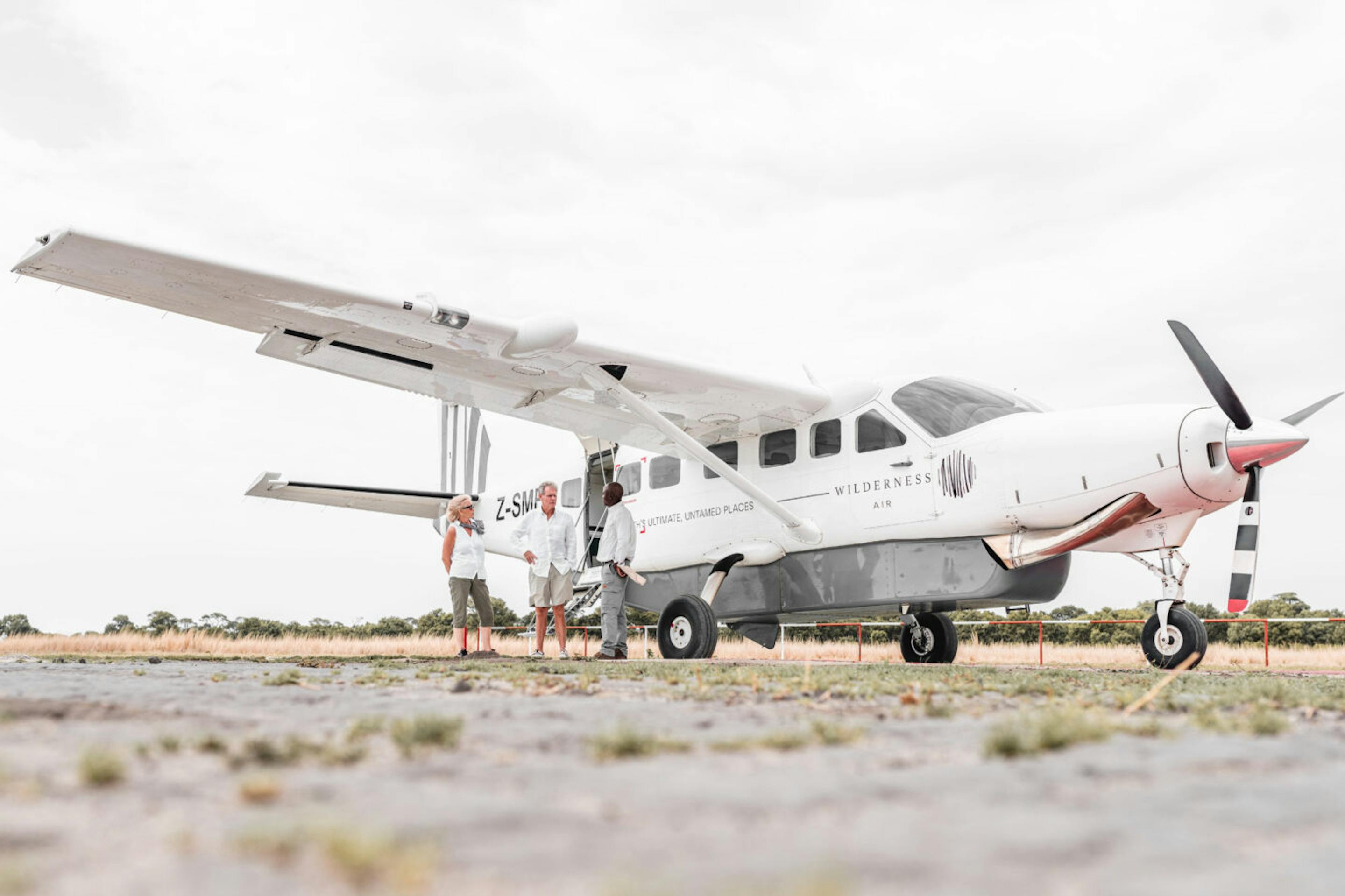 A small propeller plane is parked on a gravel airstrip, its white fuselage set against a wide gray sky overhead.