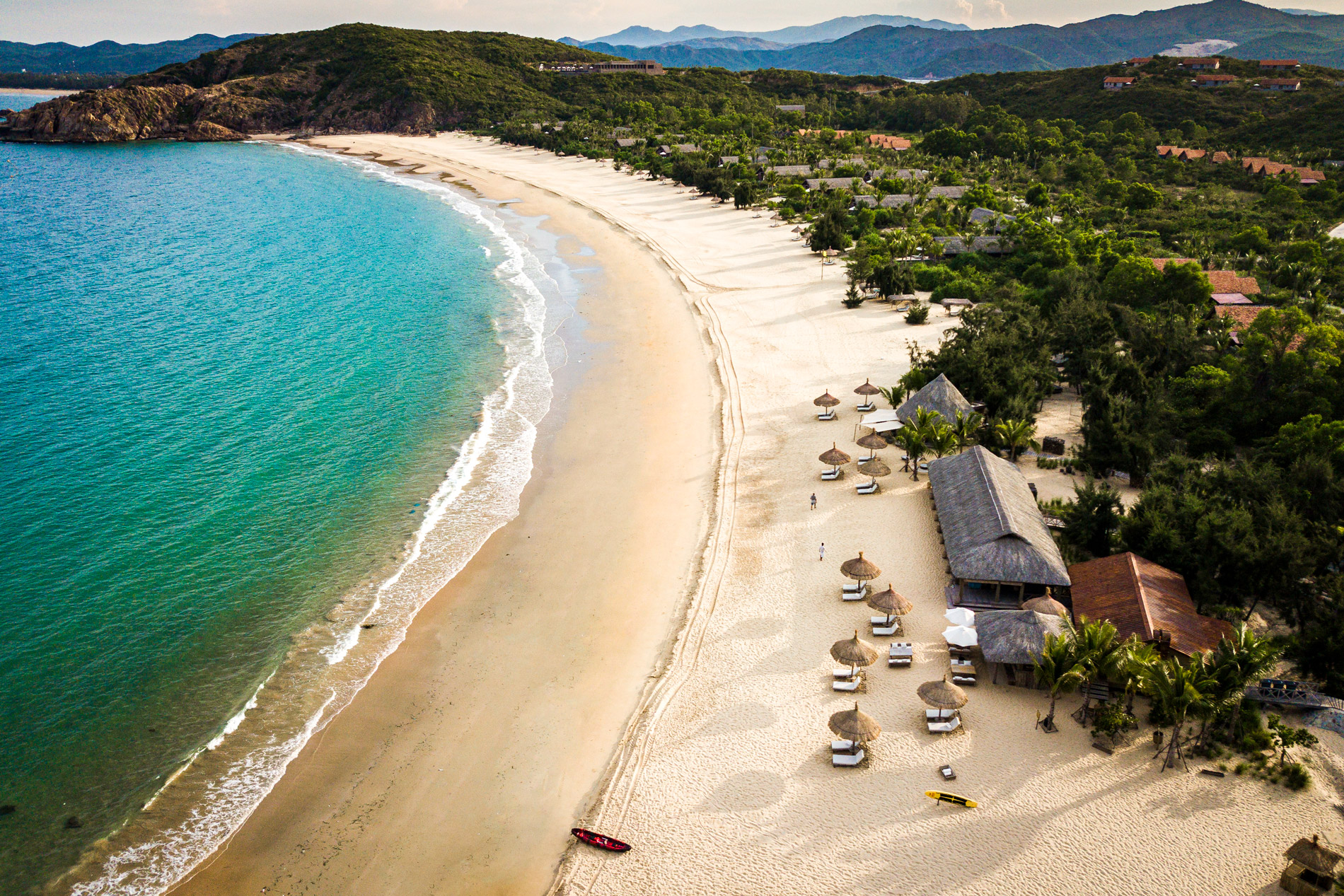 An aerial view shows a curved sandy beach and turquoise bay, with forested hills and villas set back from the shore.