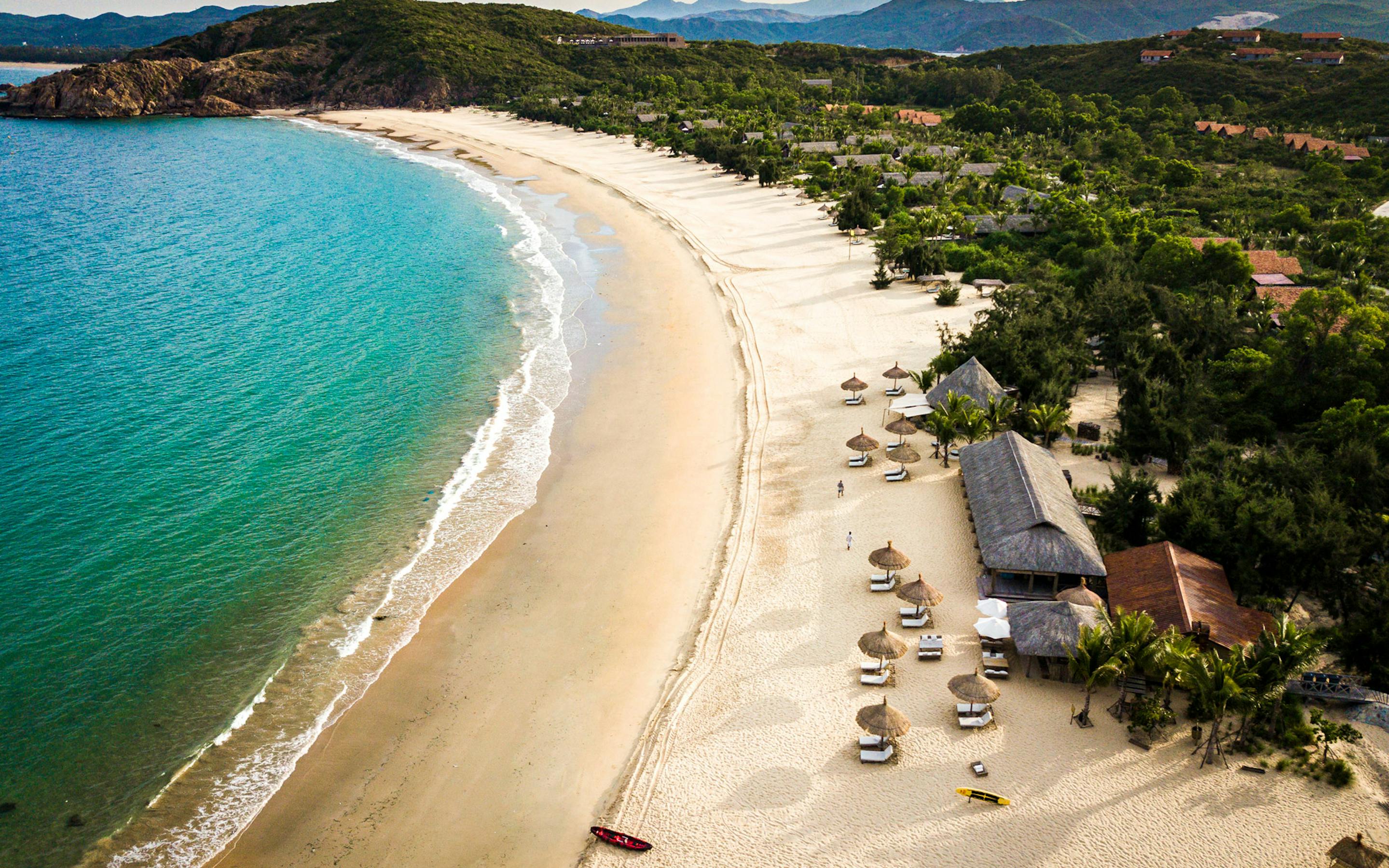 An aerial view shows a curved sandy beach and turquoise bay, with forested hills and villas set back from the shore.