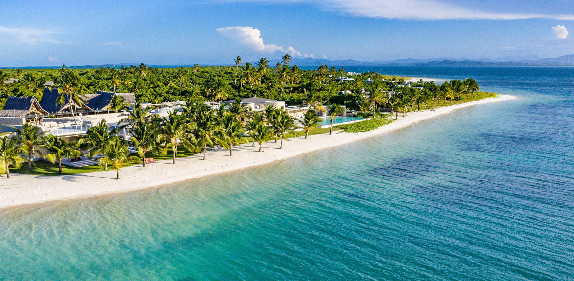 Aerial view of a tropical island resort with palm-lined beach and turquoise water fading into deep blue sea.