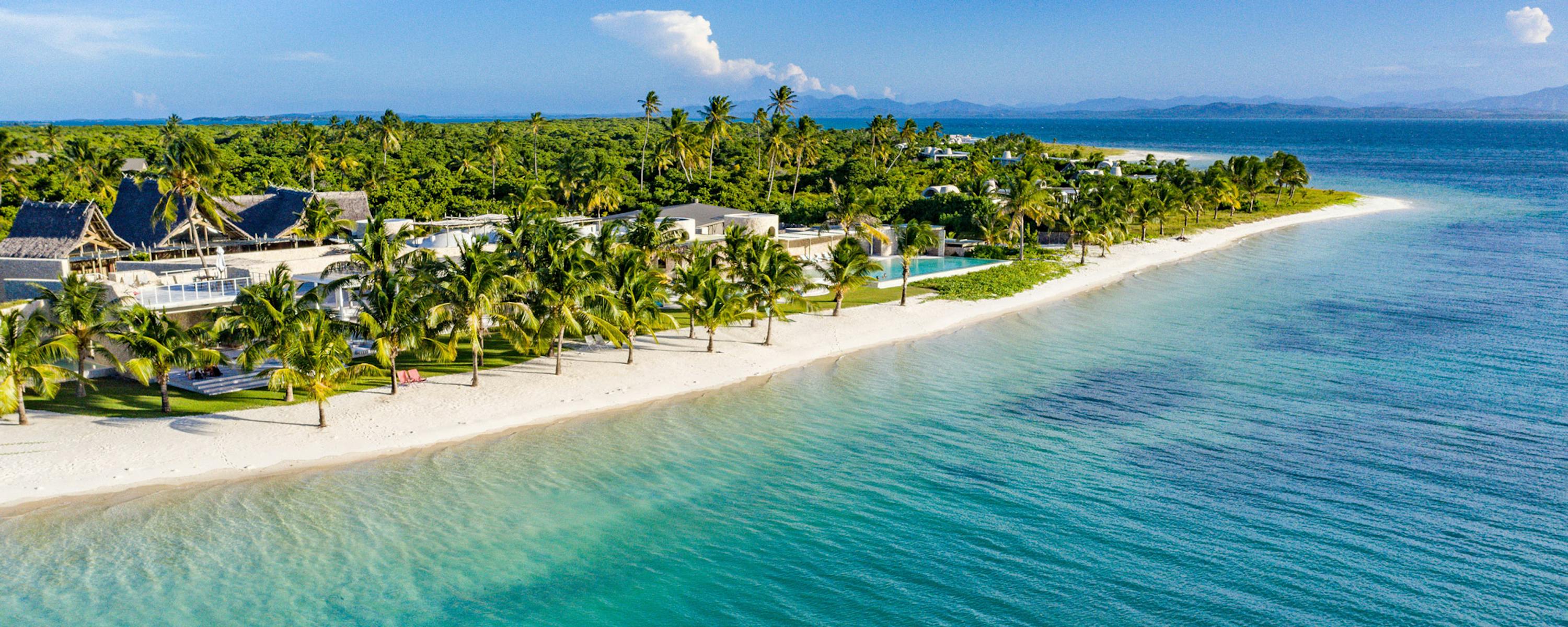 Aerial view of a tropical island resort with palm-lined beach and turquoise water fading into deep blue sea.