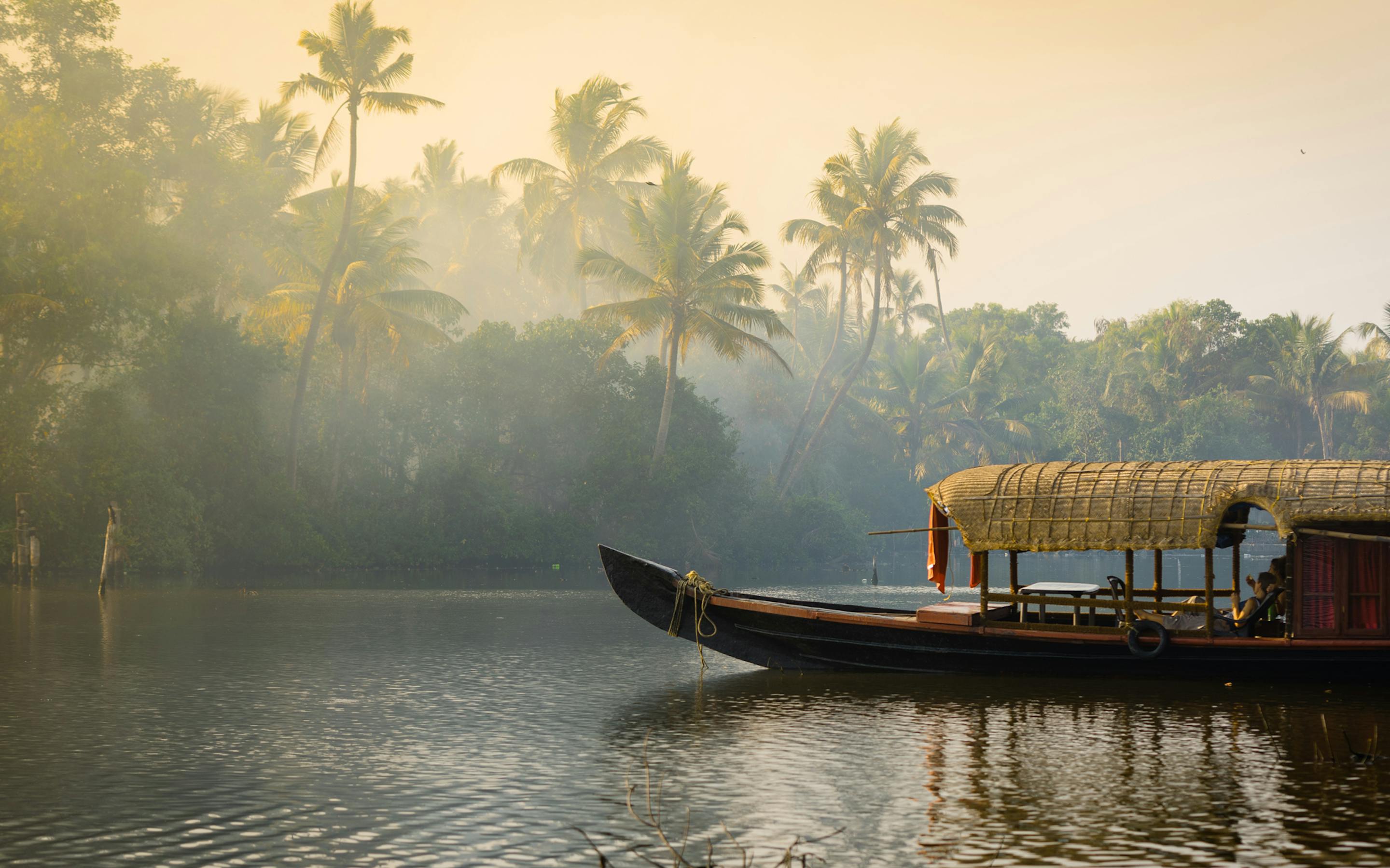 A traditional boat glides along Kerala backwaters at dawn, with palm trees and mist rising above the calm waterway.