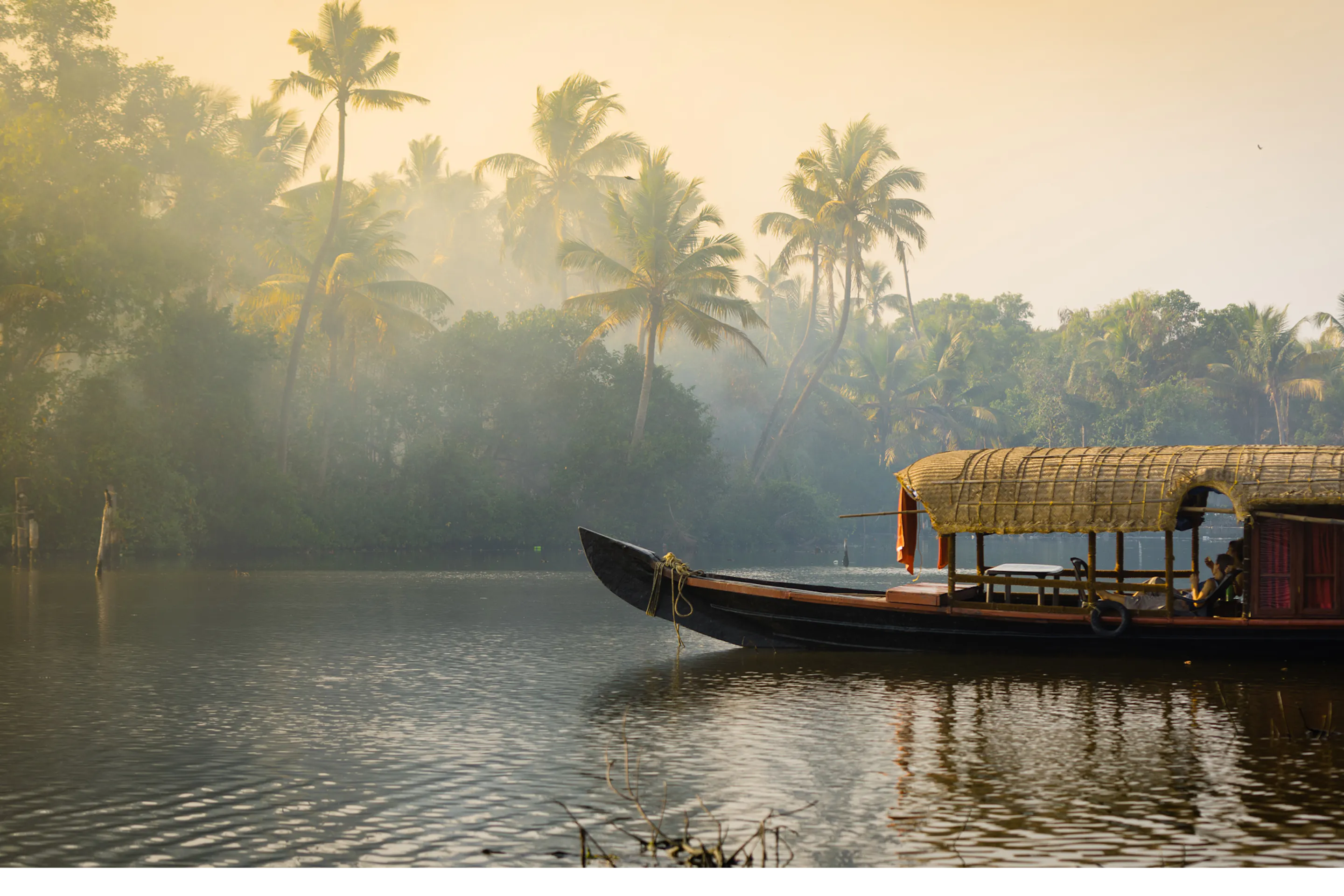 A traditional boat glides along Kerala backwaters at dawn, with palm trees and mist rising above the calm waterway.
