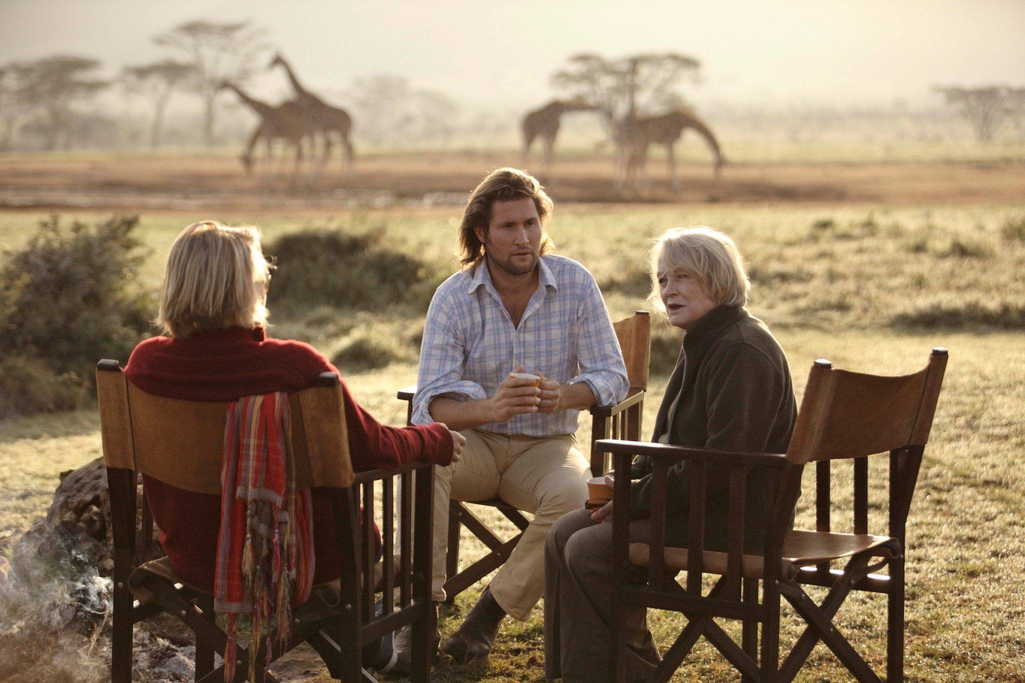 Three people sit at an outdoor table in tall grass while giraffes walk across the savanna in the hazy background.