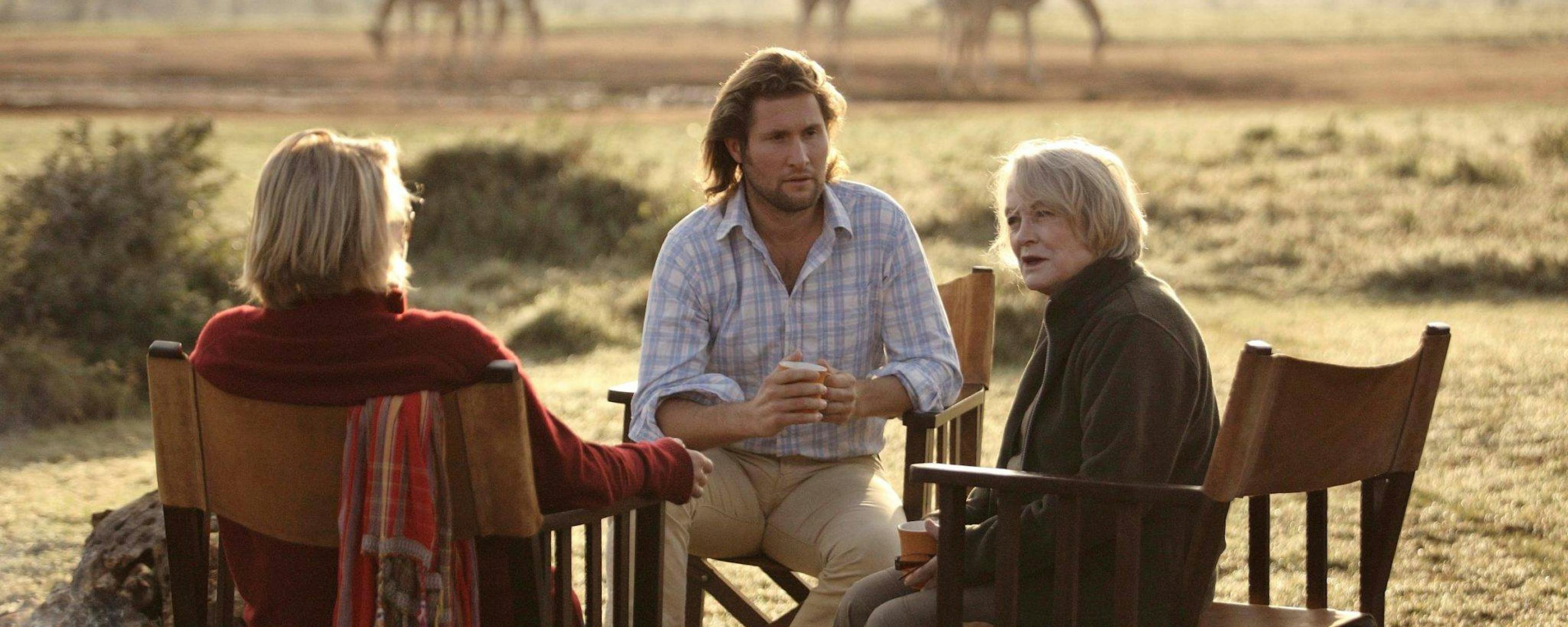 Three people sit at an outdoor table in tall grass while giraffes walk across the savanna in the hazy background.