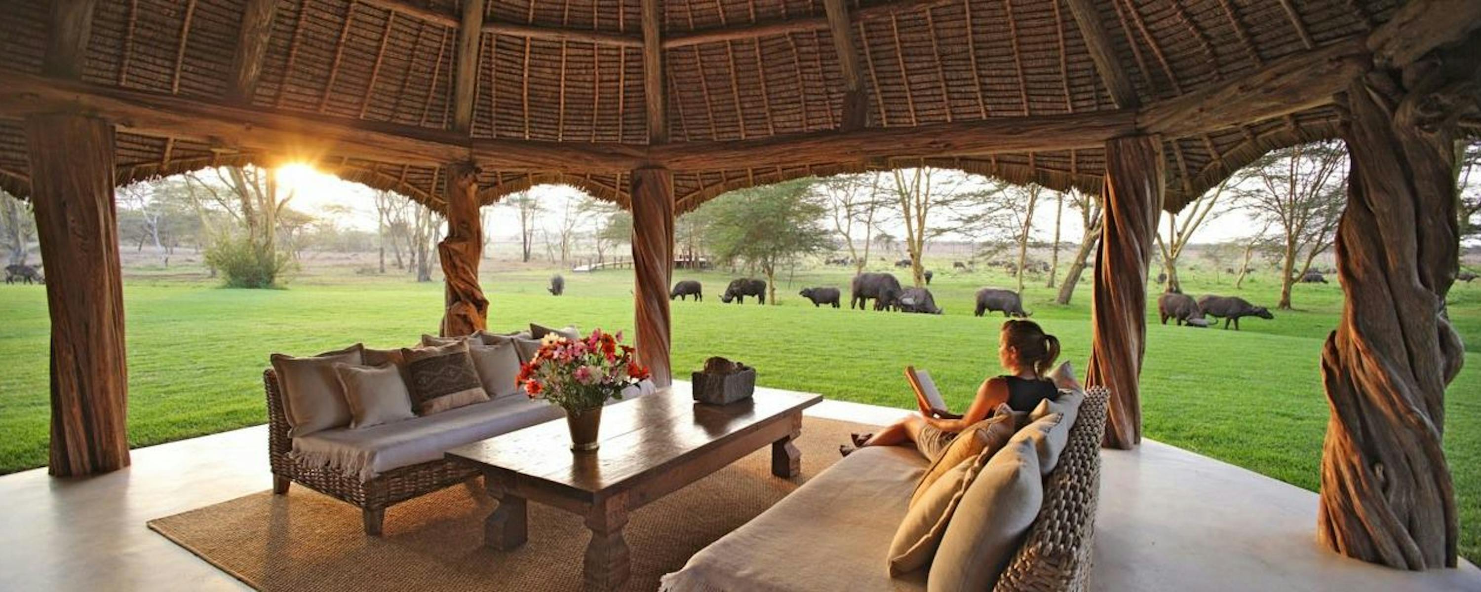 A person relaxes on cushioned lounge chairs under a thatched roof, overlooking green lawn and scattered trees.