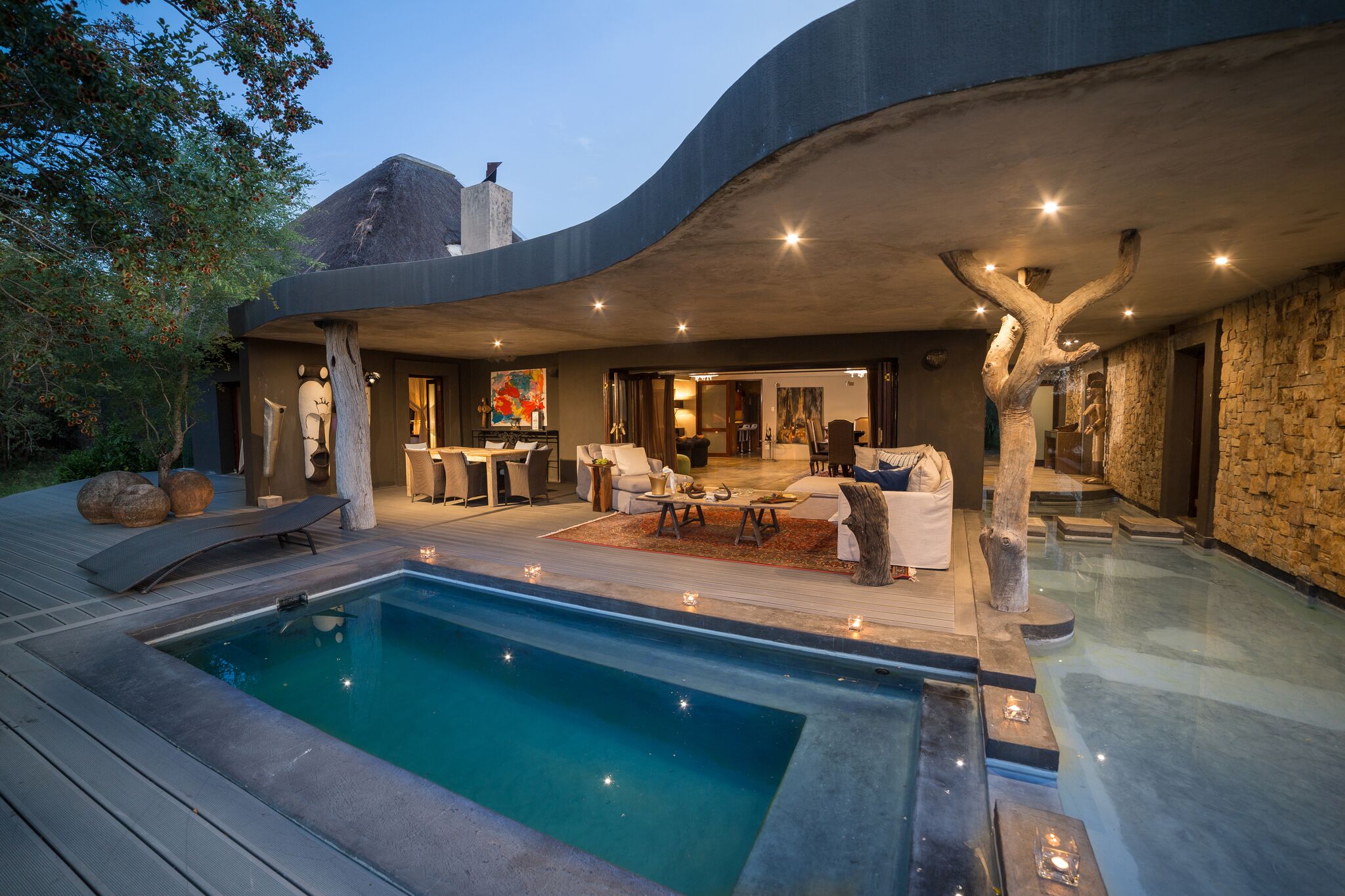 A modern lodge courtyard glows at dusk beside a turquoise pool, with outdoor seating and curved rooflines.