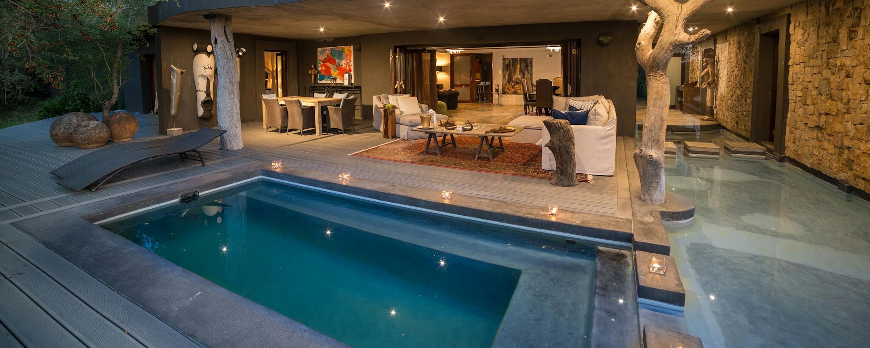 A modern lodge courtyard glows at dusk beside a turquoise pool, with outdoor seating and curved rooflines.