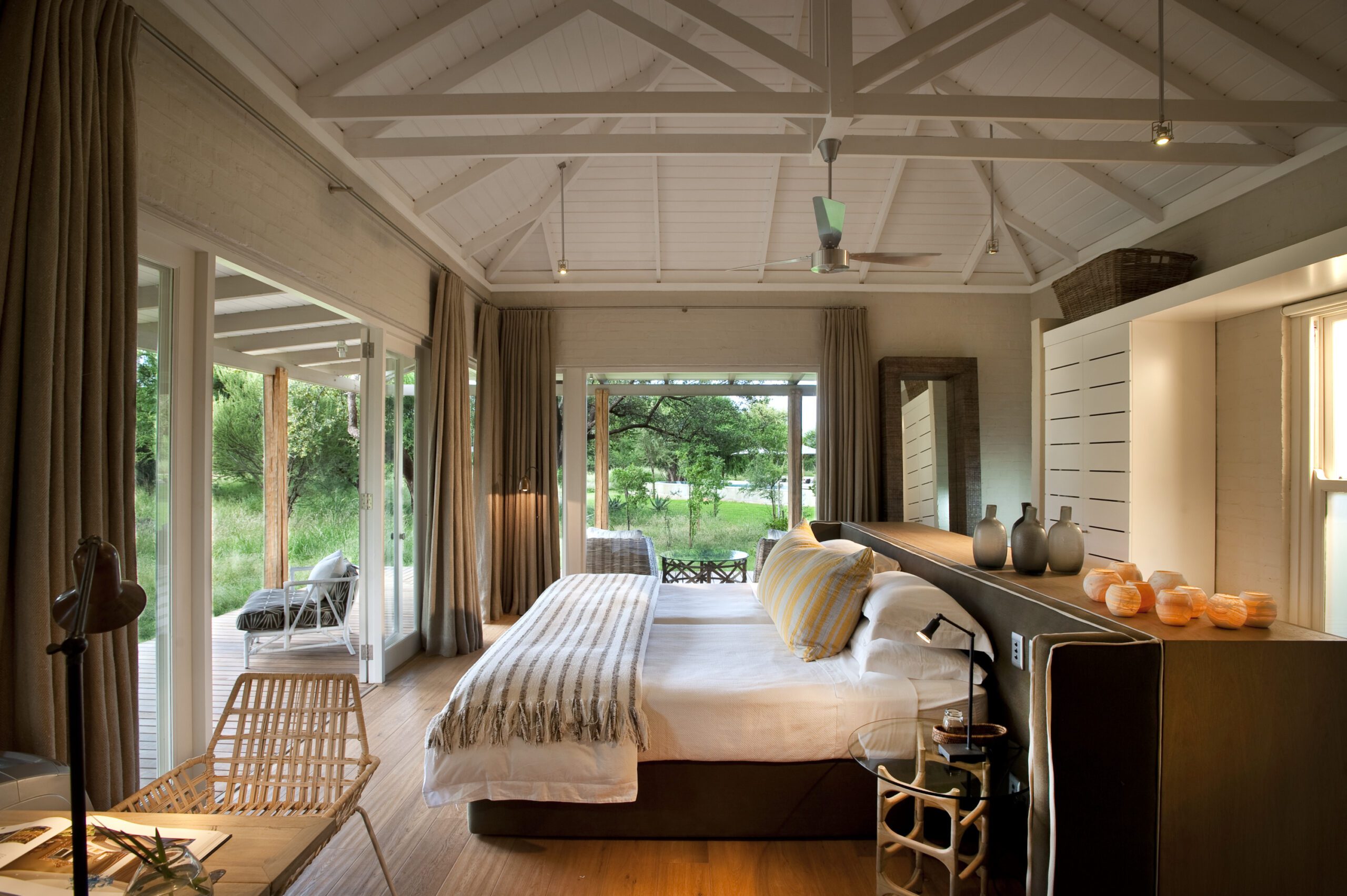 Bright bedroom with twin beds and white rafters opens to garden views through wide glass doors and soft curtains.