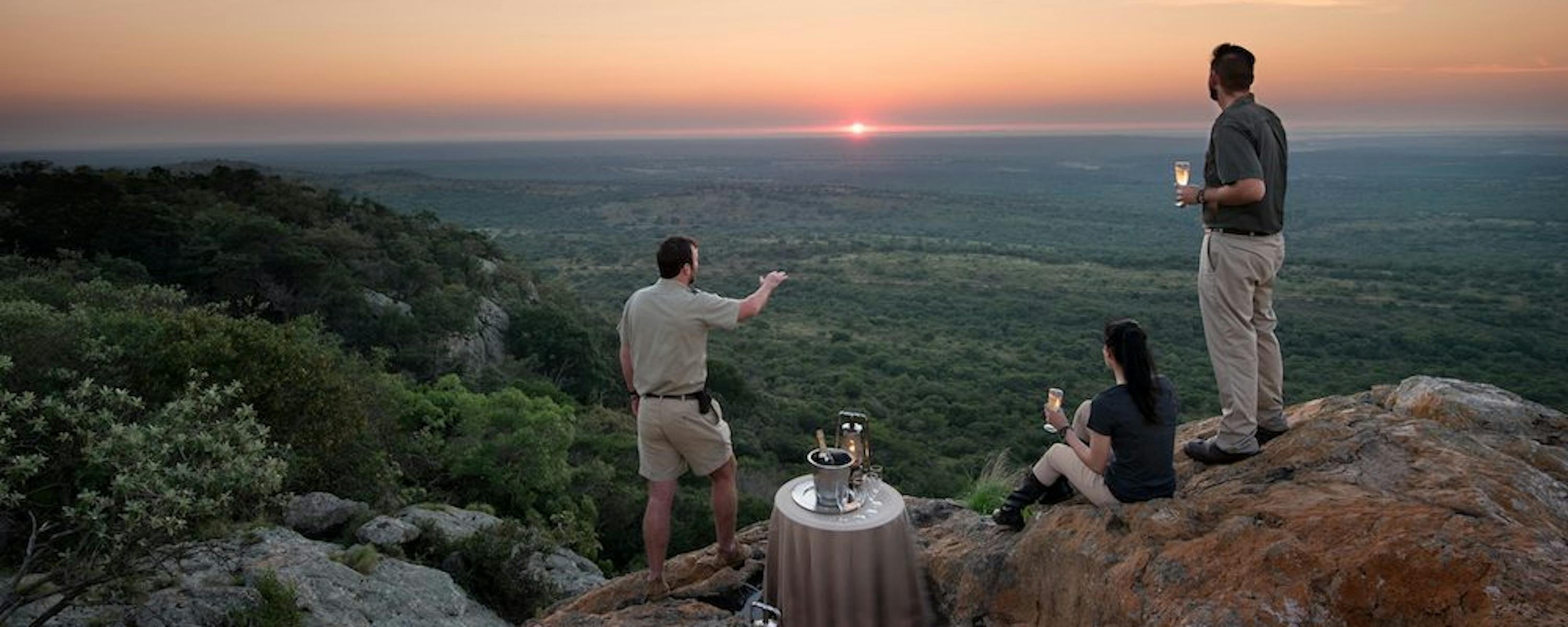 Three people gather by a small table on a rocky lookout as the sun sets, revealing layered hills and forest below.
