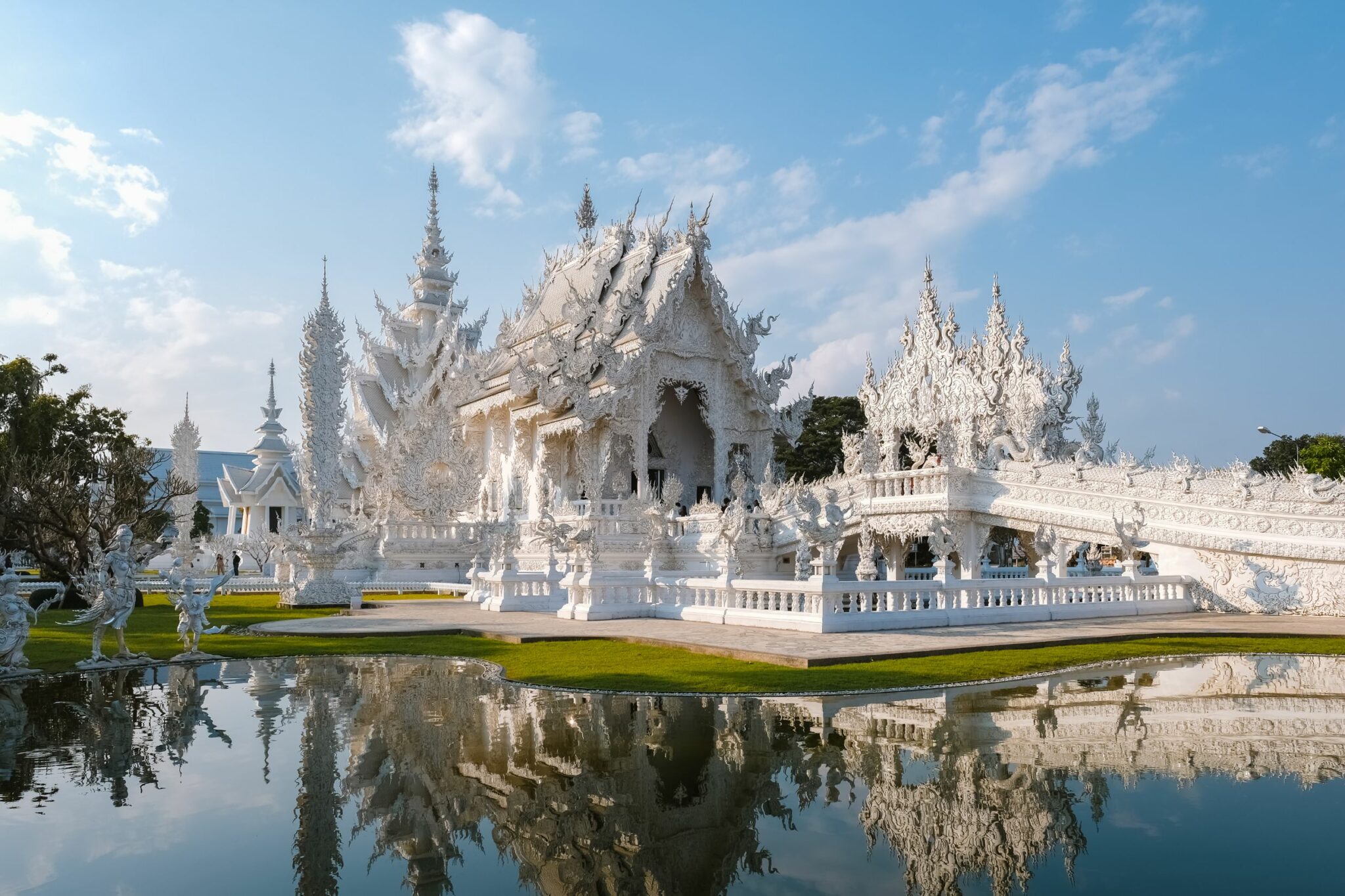 An ornate white temple reflects in a still pond, with pointed spires, mirrored water, and bright blue sky above.