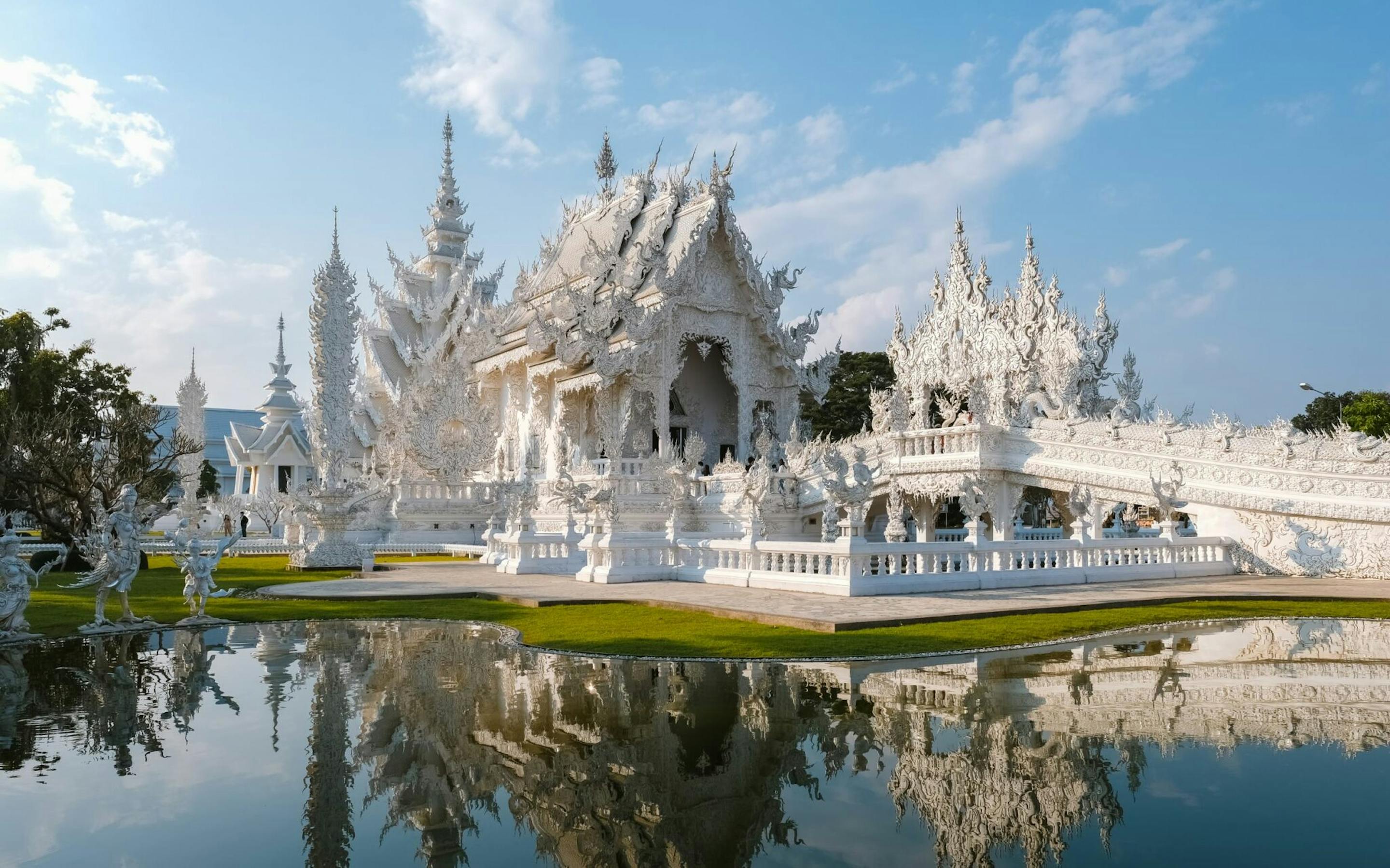 An ornate white temple reflects in a still pond, with pointed spires, mirrored water, and bright blue sky above.