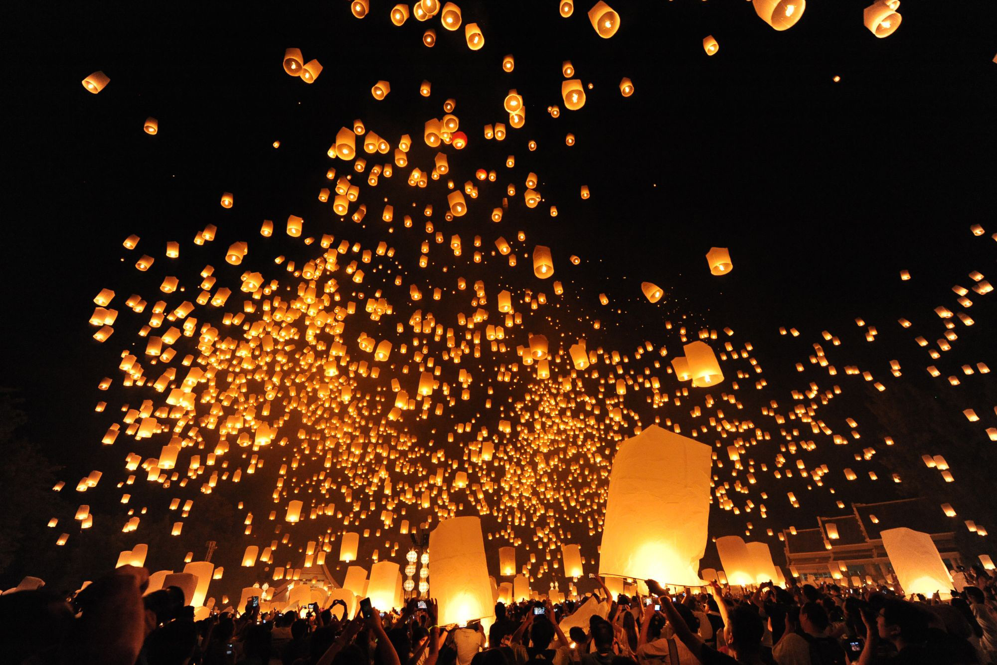 Hundreds of glowing sky lanterns rise above a nighttime crowd, scattering warm light against a black sky.