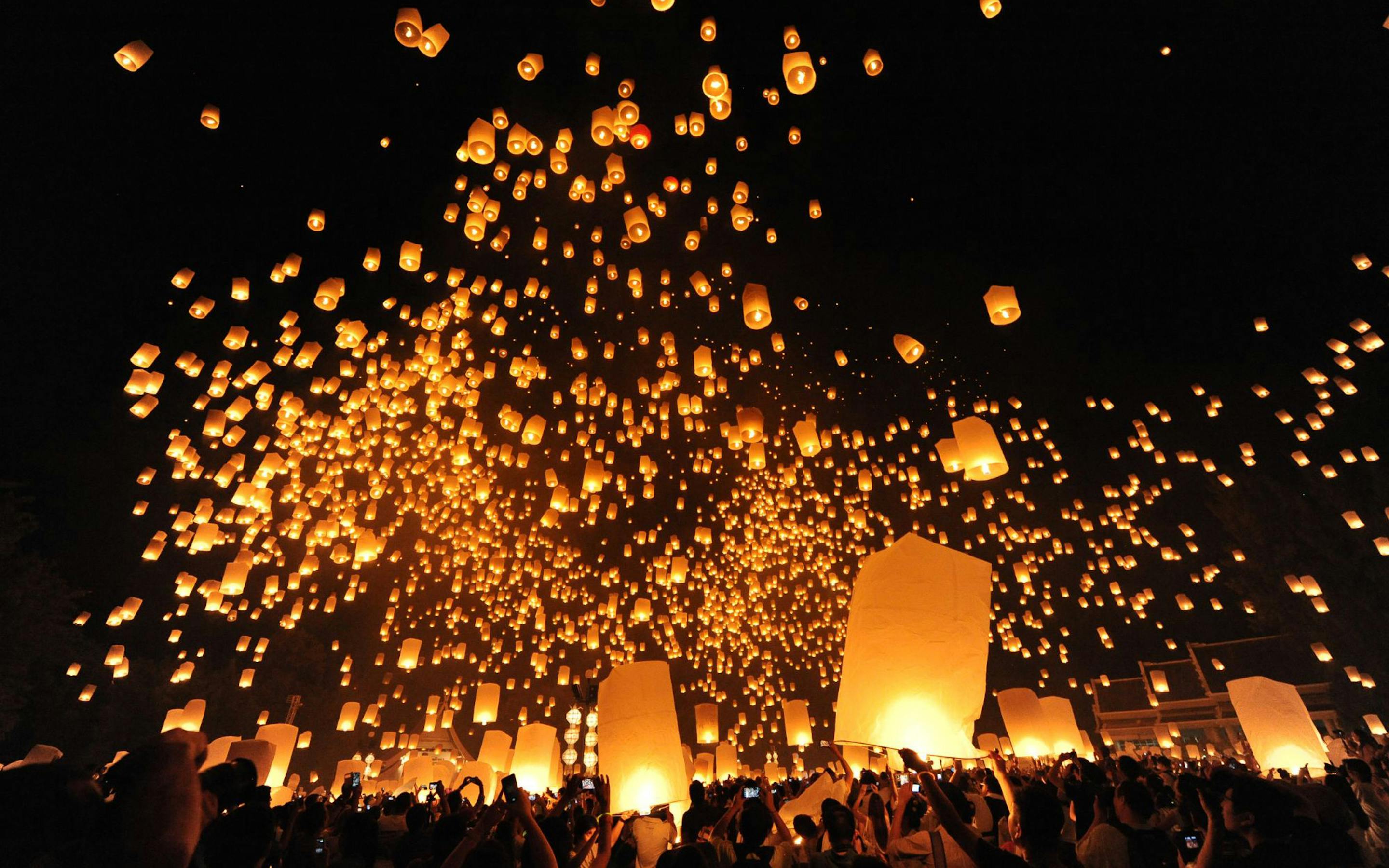 Hundreds of glowing sky lanterns rise above a nighttime crowd, scattering warm light against a black sky.