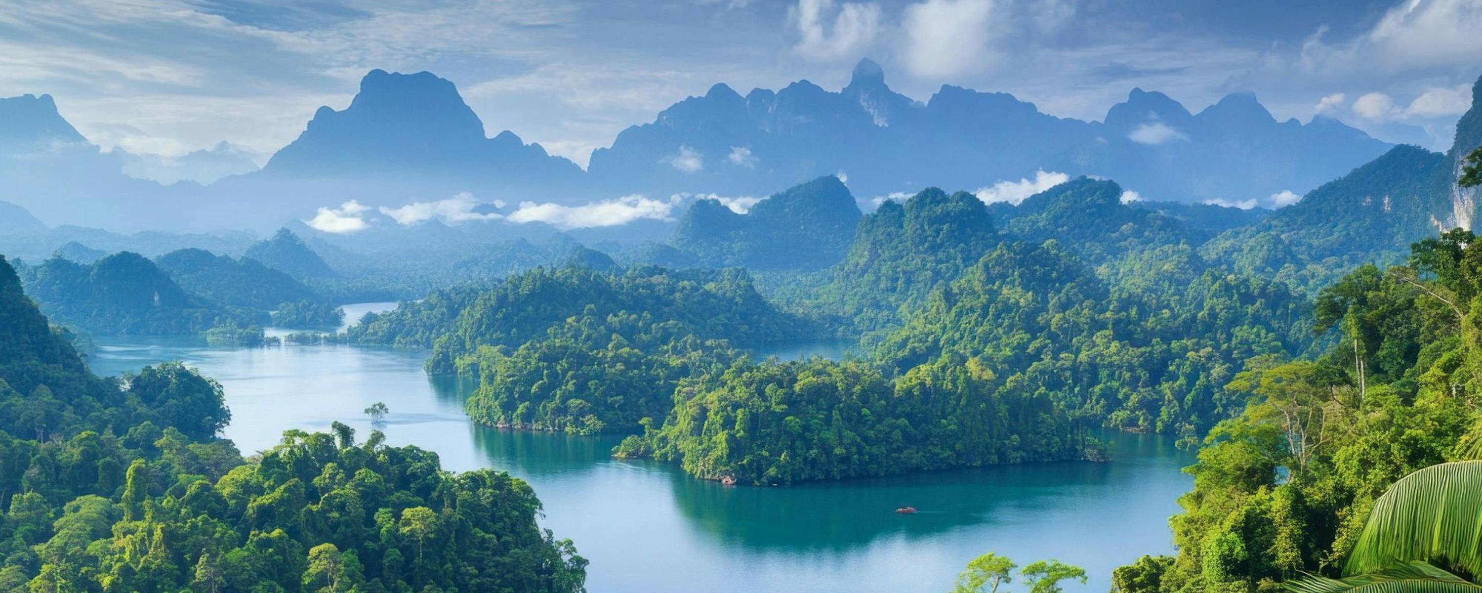 An emerald lake winds through dense jungle, with limestone peaks rising beyond layered, misty ridgelines in the distance.