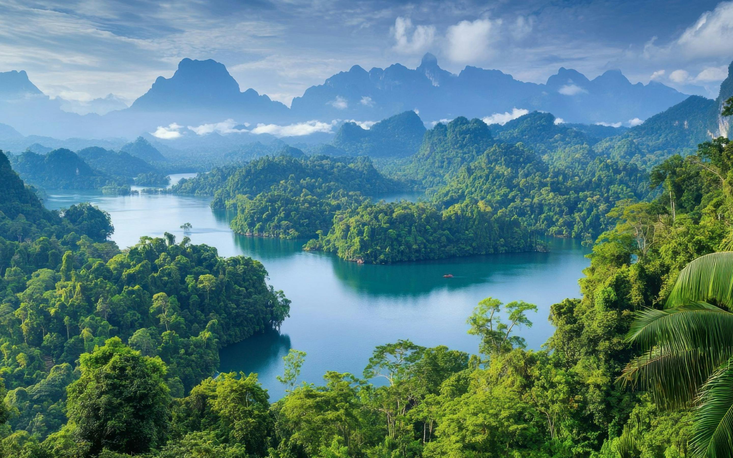 An emerald lake winds through dense jungle, with limestone peaks rising beyond layered, misty ridgelines in the distance.