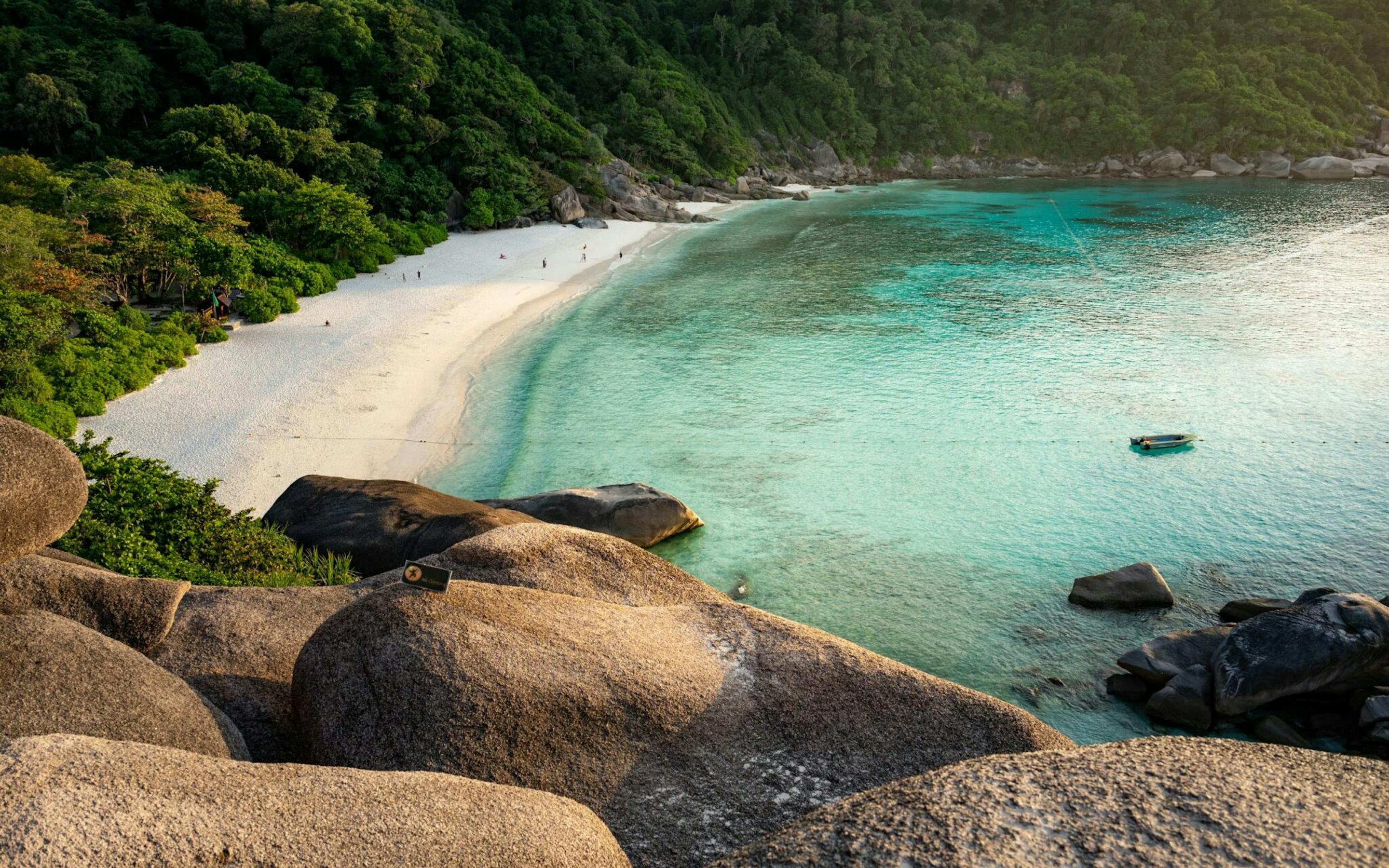 Turquoise water laps a crescent beach below smooth granite boulders, with forested hills framing the bay.