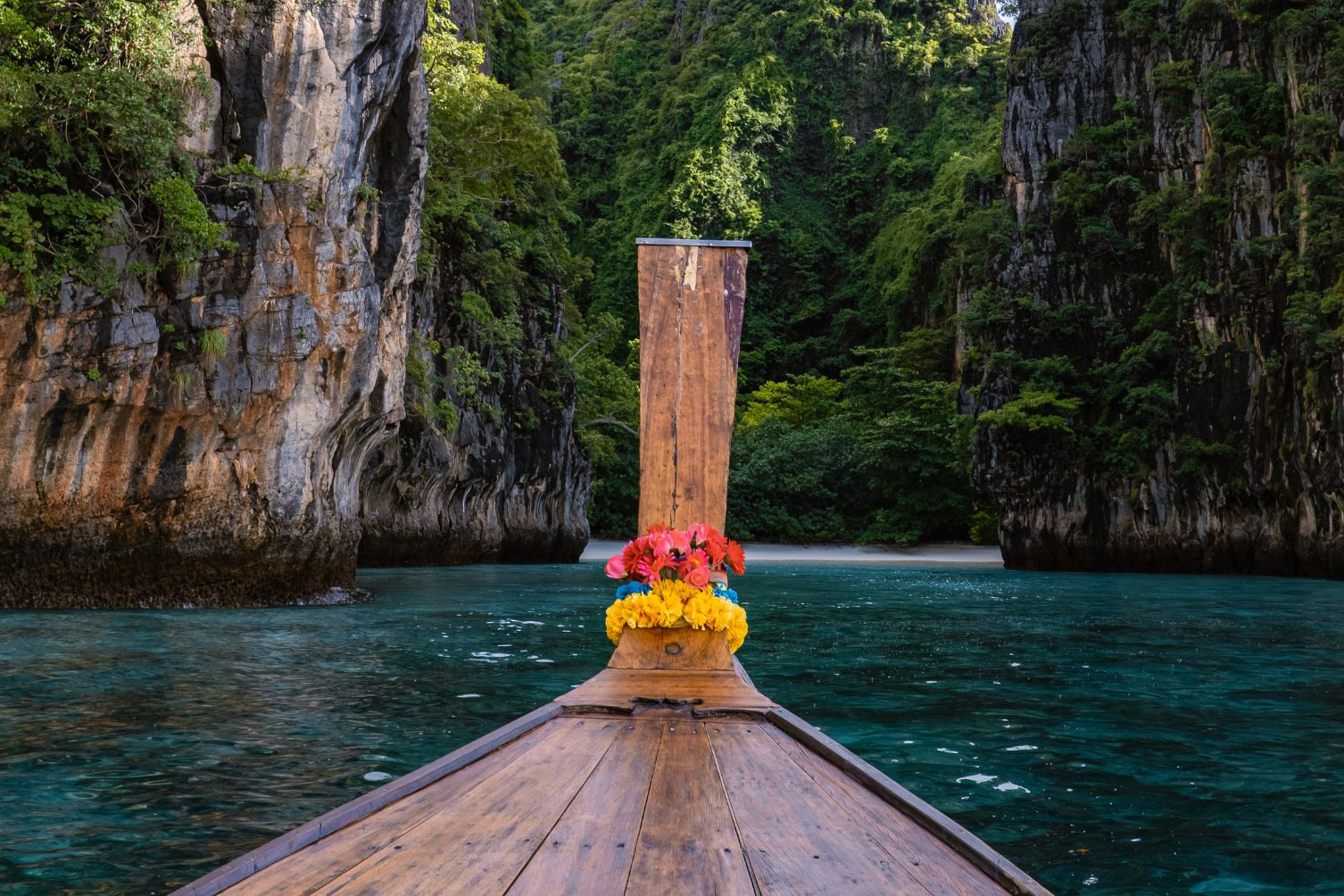 A longtail boat points into a narrow lagoon, with steep limestone cliffs, vines, and dark green water ahead.
