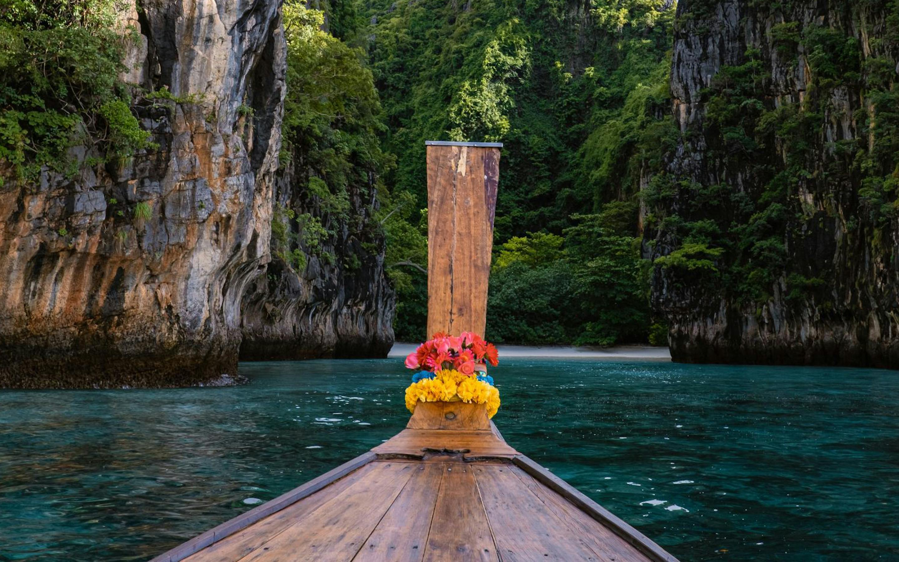A longtail boat points into a narrow lagoon, with steep limestone cliffs, vines, and dark green water ahead.