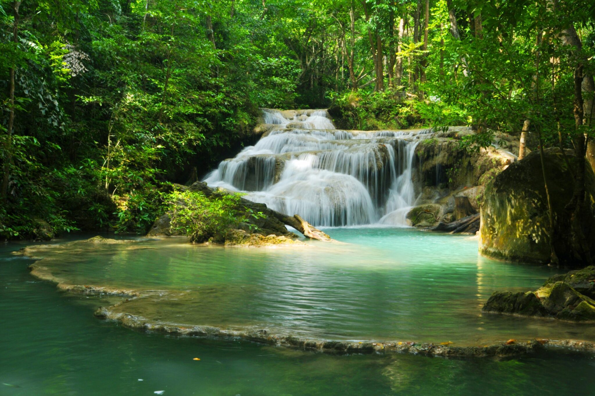 Tiered waterfalls spill into jade pools in a lush forest, with sunlight filtering through leaves and mossy rocks.