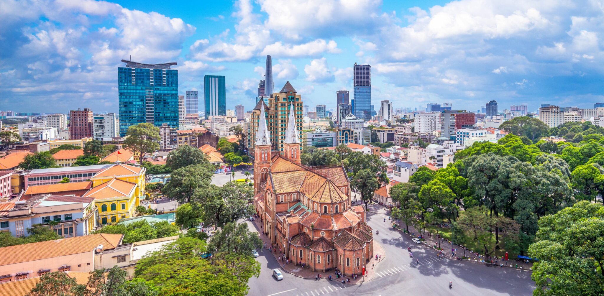 Aerial view of Ho Chi Minh City with a red-brick cathedral, leafy streets, and modern towers under clouds.