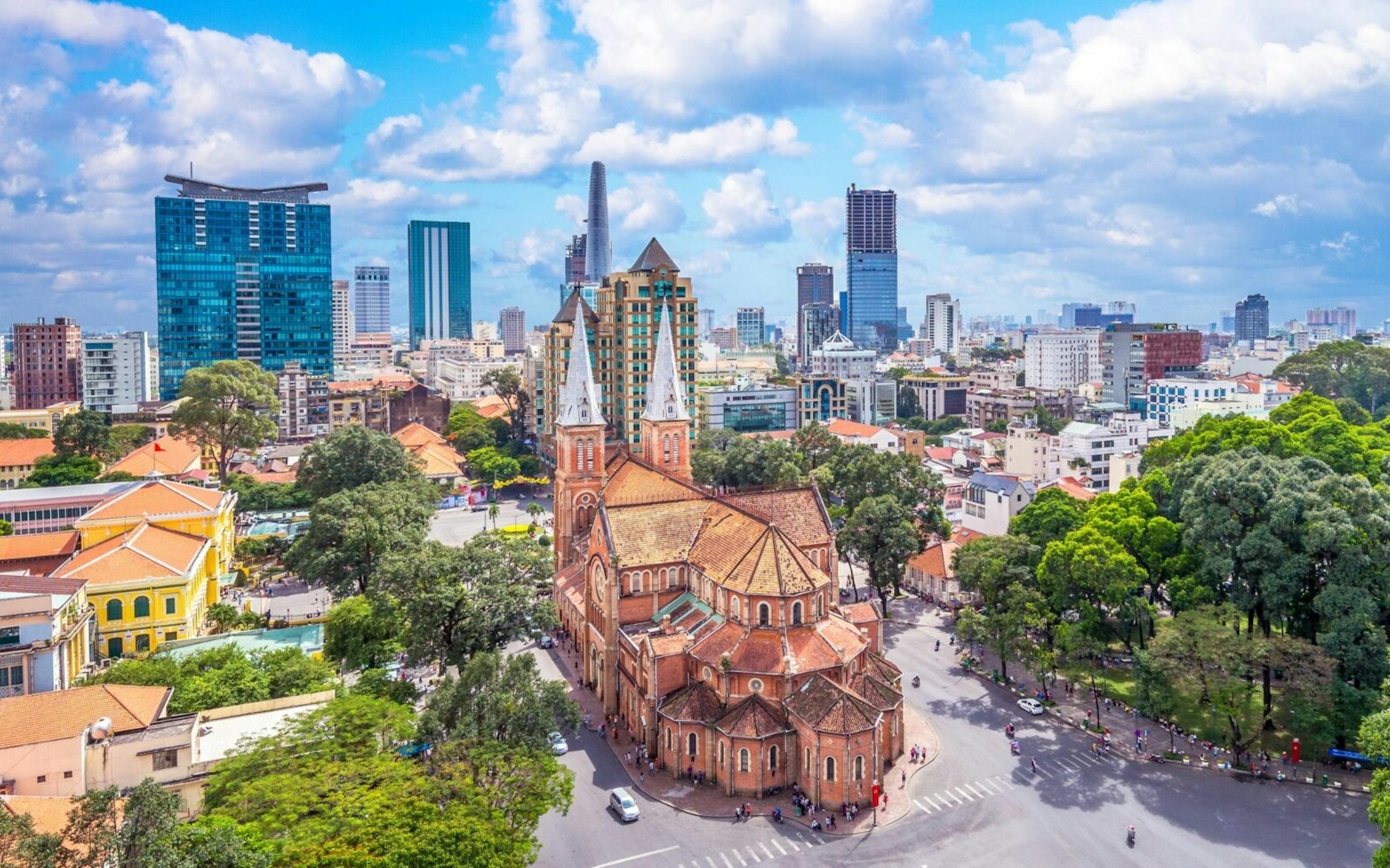 Aerial view of Ho Chi Minh City with a red-brick cathedral, leafy streets, and modern towers under clouds.