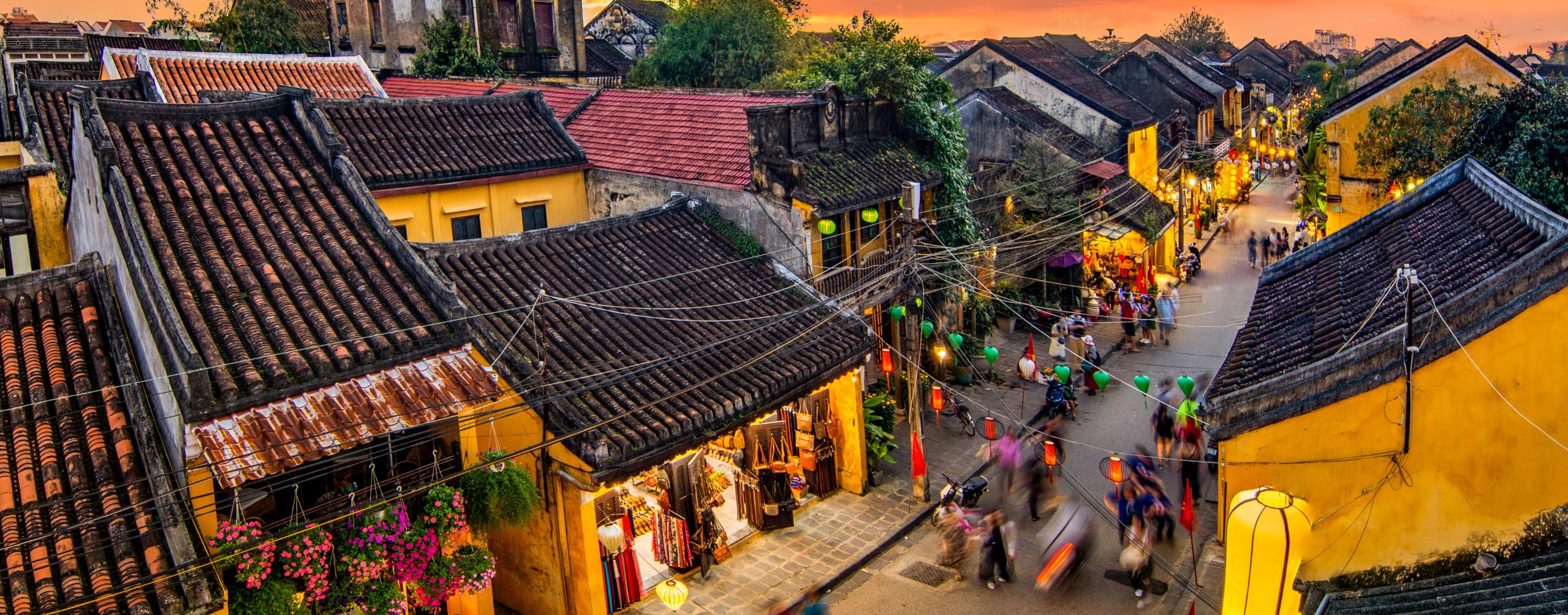 A bustling street in Hoi An is lined with yellow buildings and lanterns, as crowds move through warm evening light.