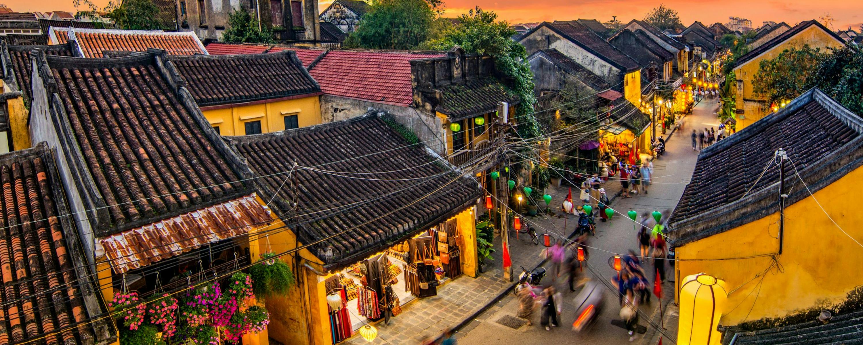 A bustling street in Hoi An is lined with yellow buildings and lanterns, as crowds move through warm evening light.