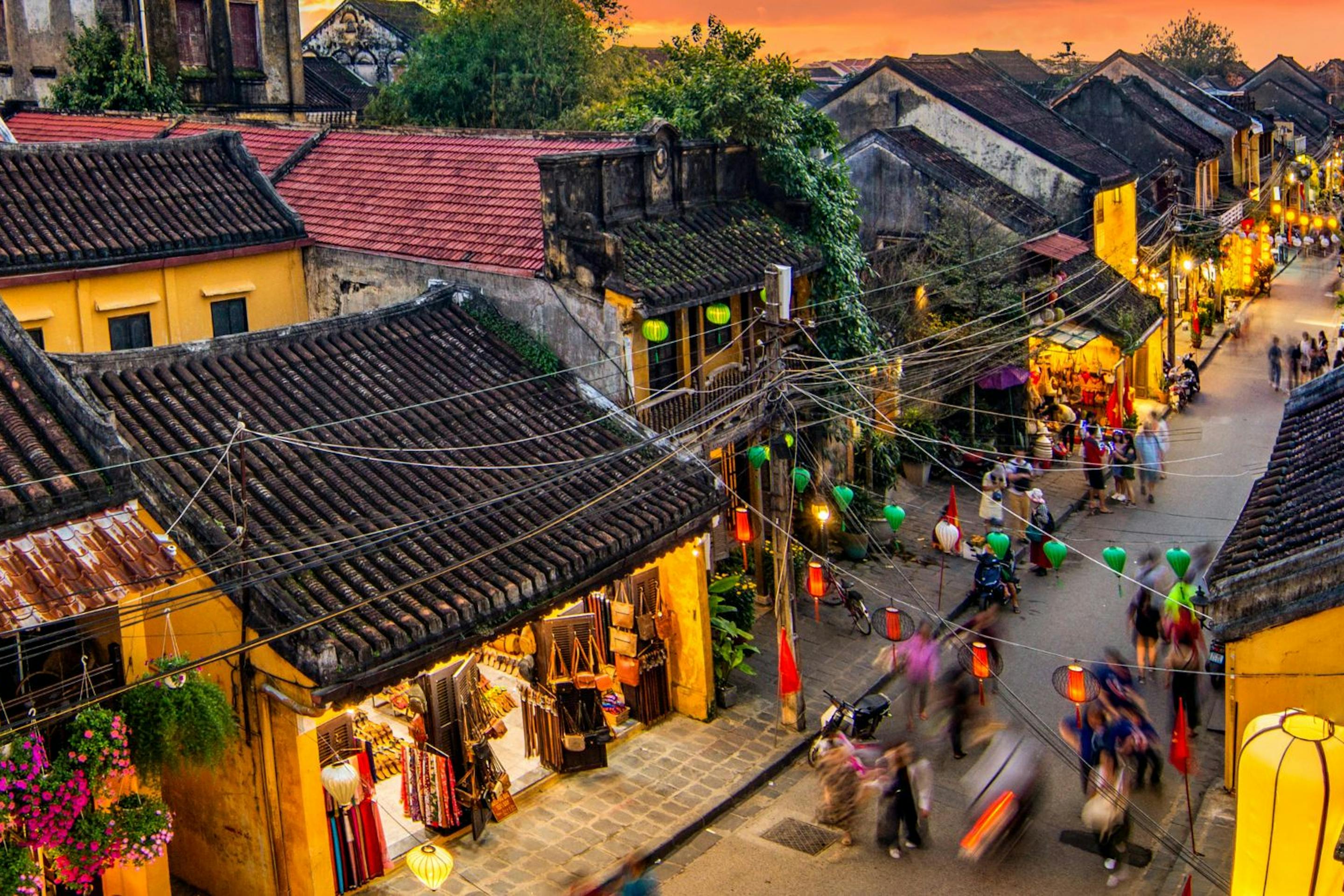 A bustling street in Hoi An is lined with yellow buildings and lanterns, as crowds move through warm evening light.