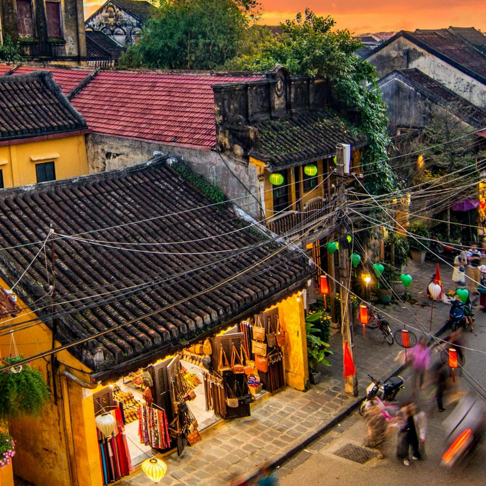 A bustling street in Hoi An is lined with yellow buildings and lanterns, as crowds move through warm evening light.