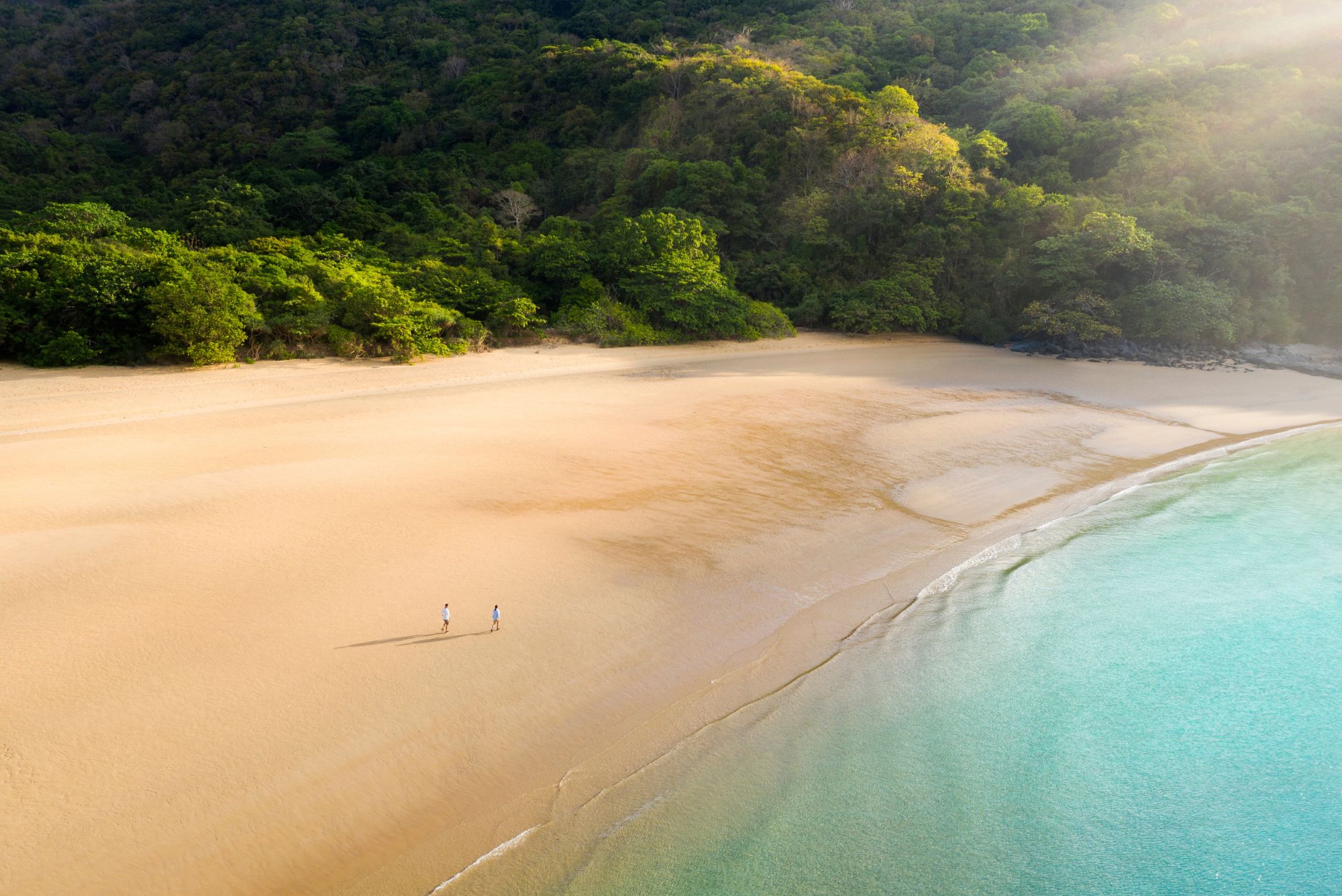 A wide sandy beach curves beside turquoise water as two people stroll along the quiet shore below green hills.