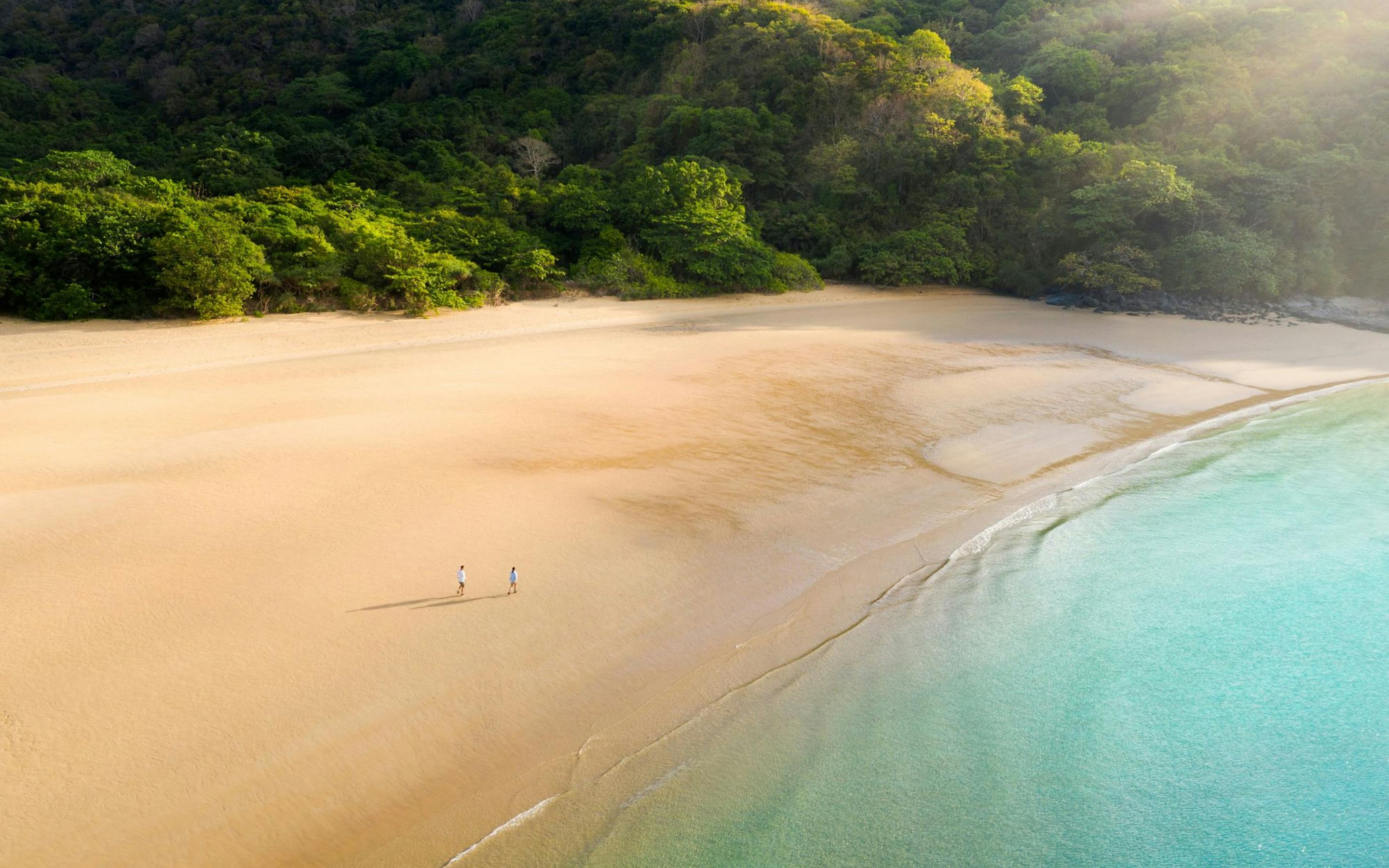 A wide sandy beach curves beside turquoise water as two people stroll along the quiet shore below green hills.
