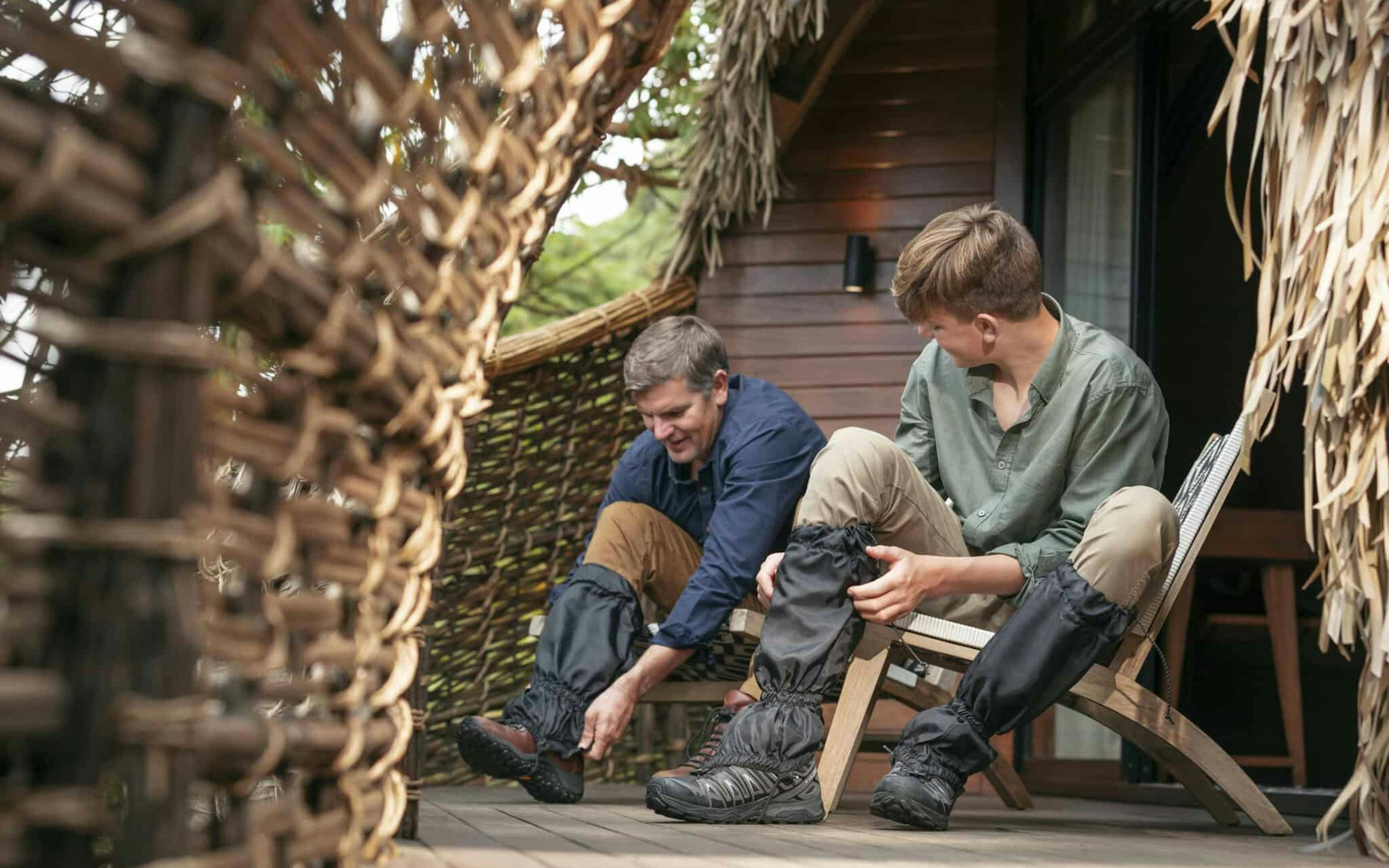 An adult and child sit on a wooden lodge deck beside woven walls, putting on boots.
