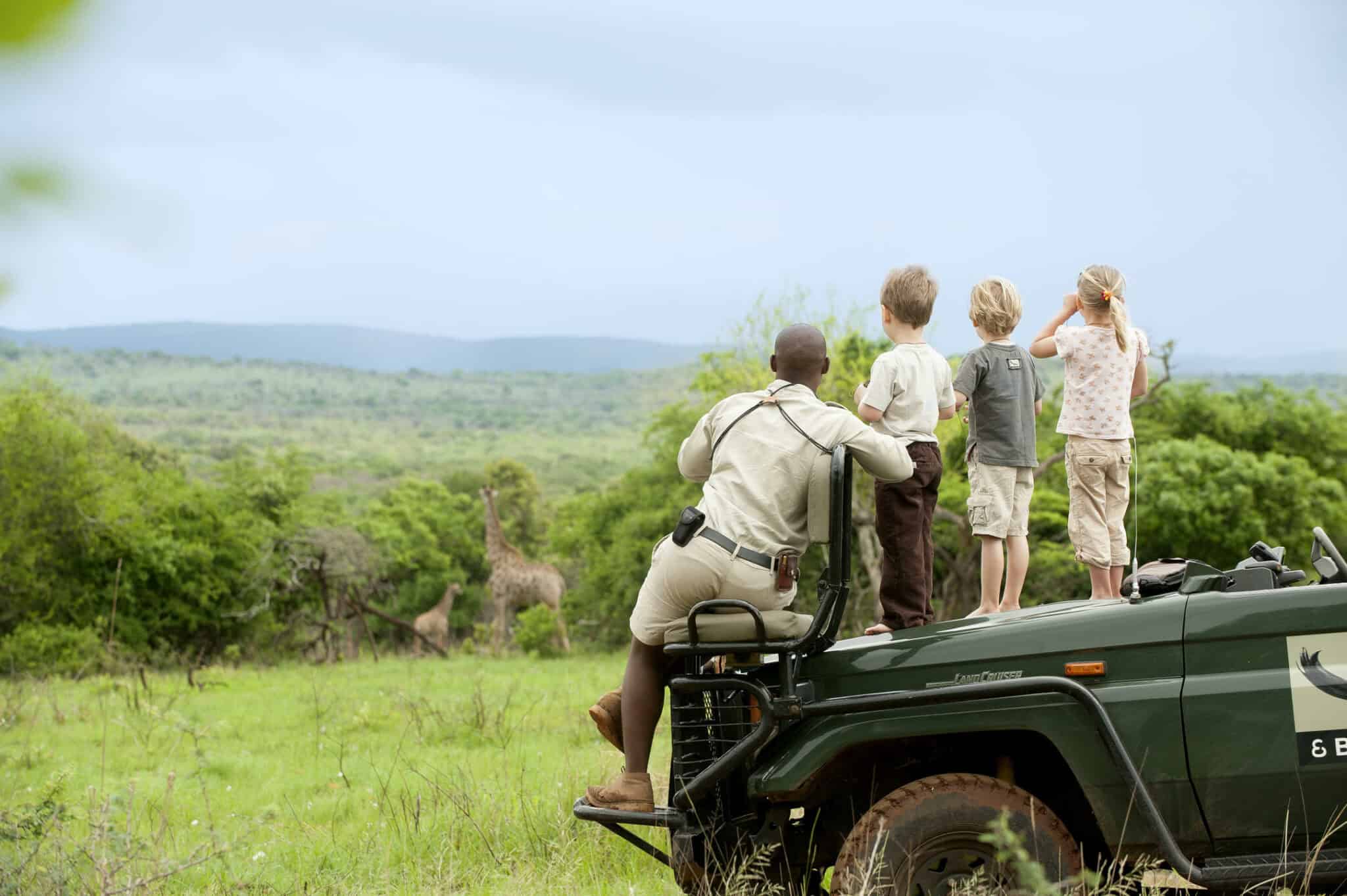 Three children and  an adults stand on a safari vehicle, watching, giraffe, with green plains stretch to the horizon under pale clouds.