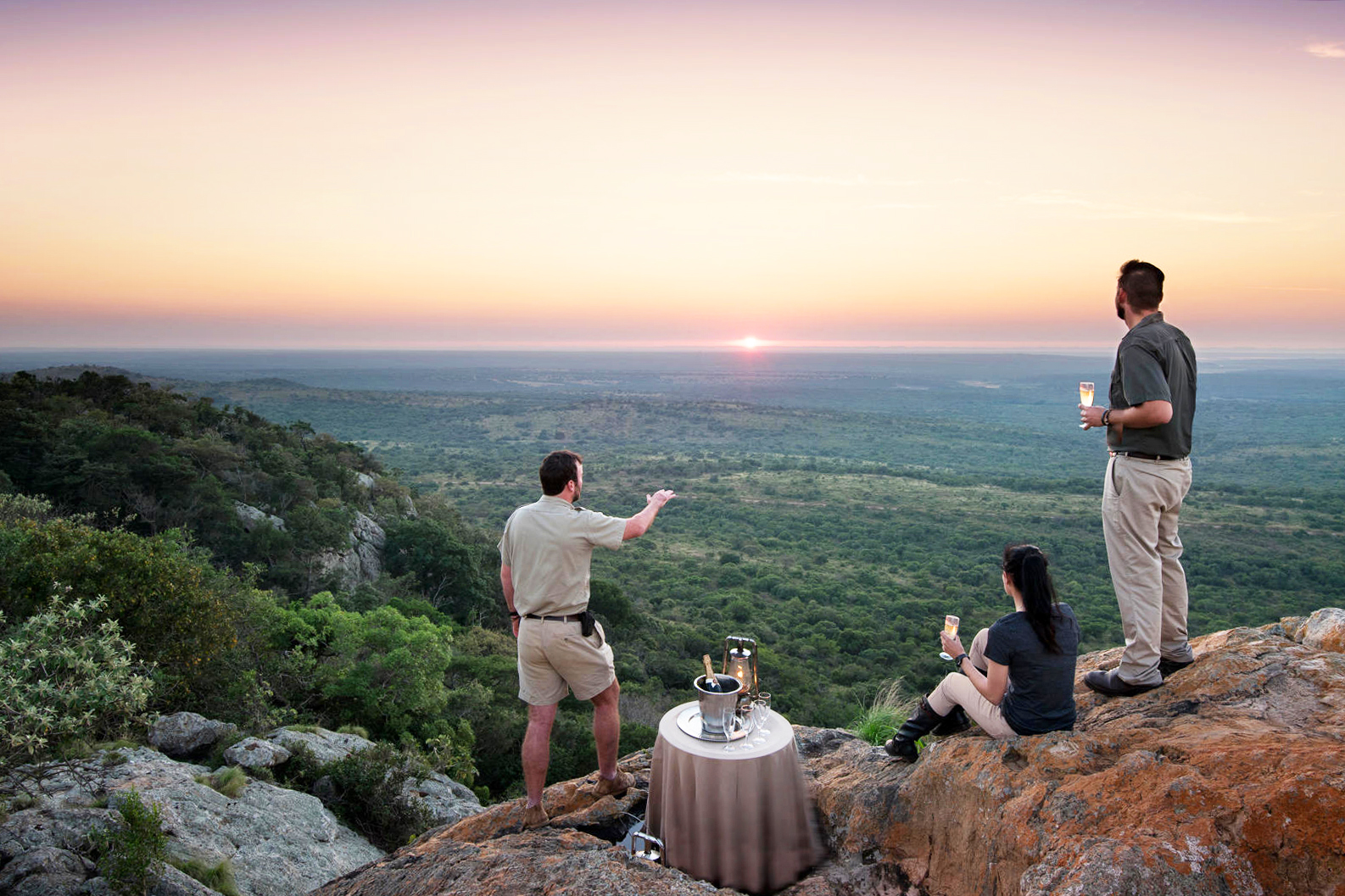 Guests gather on a rocky lookout at sunset beside drinks and lanterns, overlooking a wide green valley below.