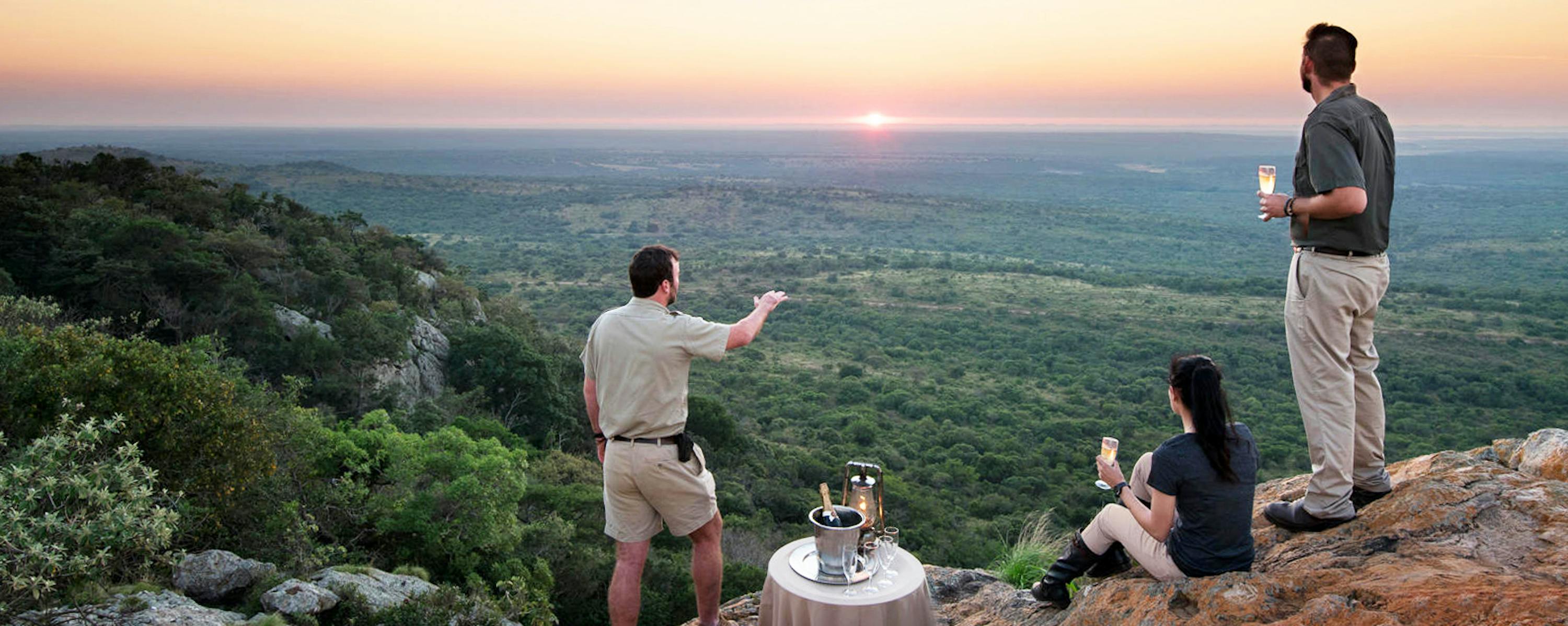 Guests gather on a rocky lookout at sunset beside drinks and lanterns, overlooking a wide green valley below.