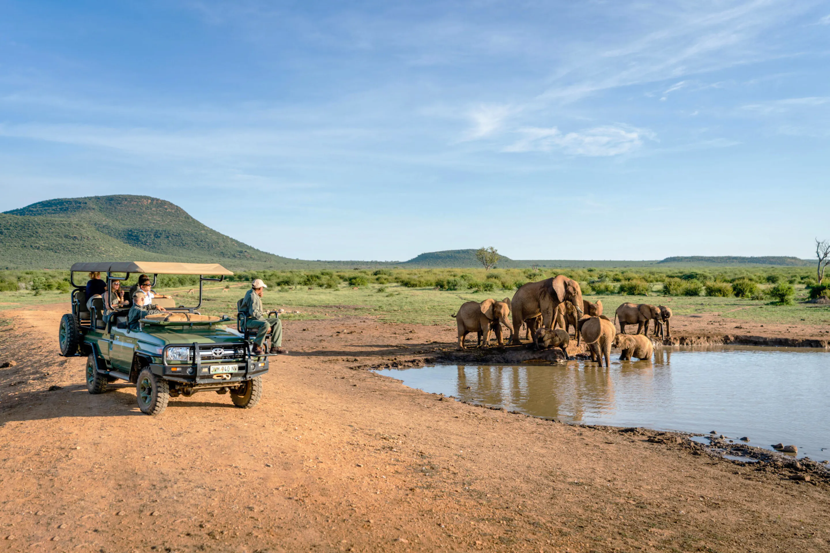 An open safari vehicle watches elephants drinking at a waterhole, with rippled reflections and green hills beyond.