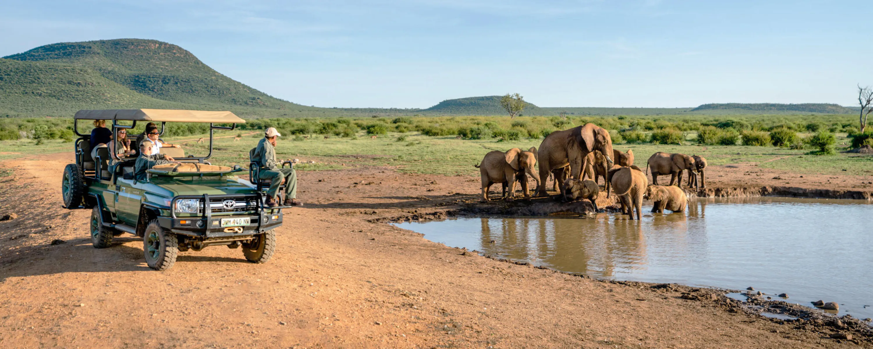 An open safari vehicle watches elephants drinking at a waterhole, with rippled reflections and green hills beyond.