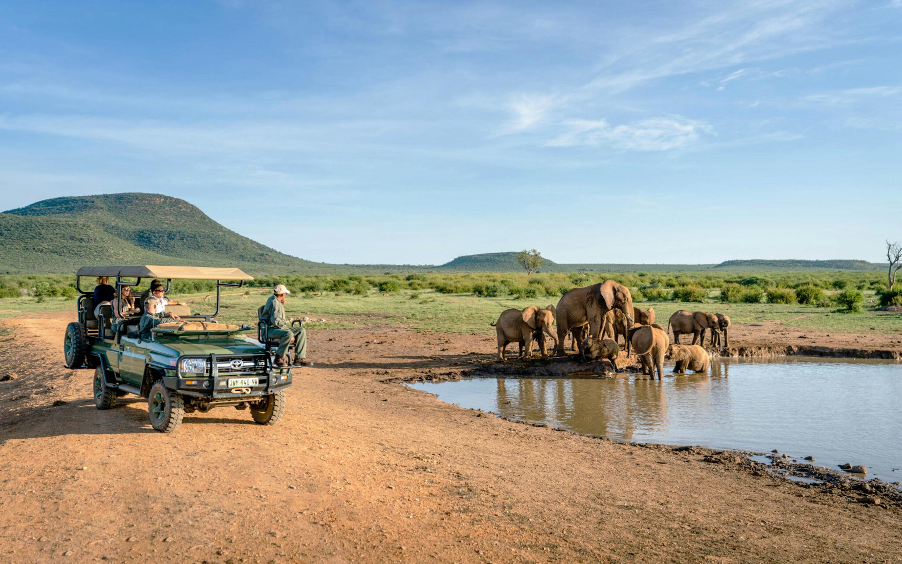 An open safari vehicle watches elephants drinking at a waterhole, with rippled reflections and green hills beyond.