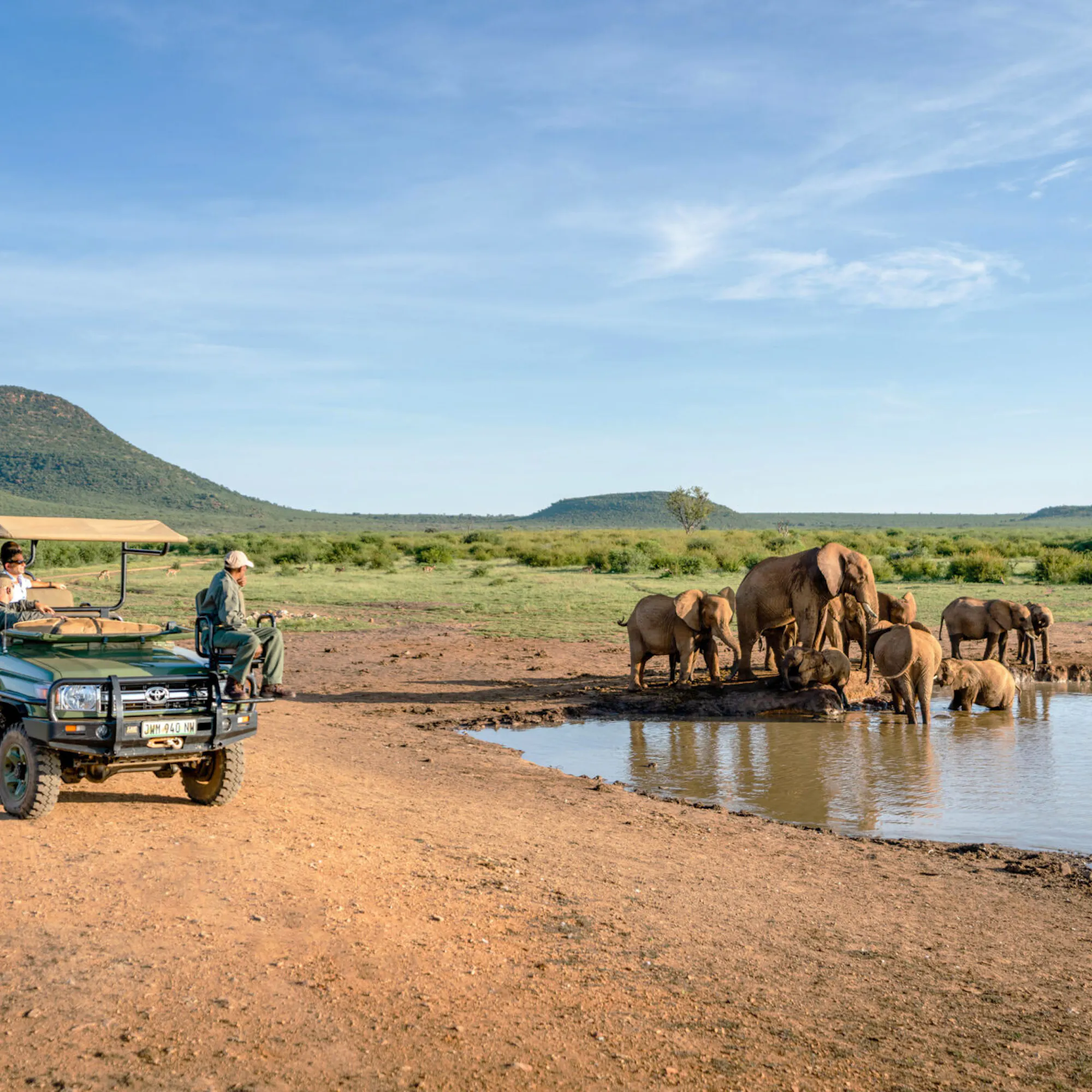 An open safari vehicle watches elephants drinking at a waterhole, with rippled reflections and green hills beyond.