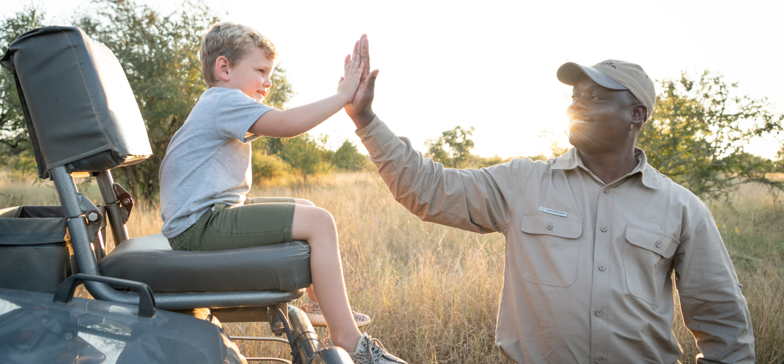 A safari guide high-fives a child seated in an open vehicle, with golden grassland and soft evening light behind.