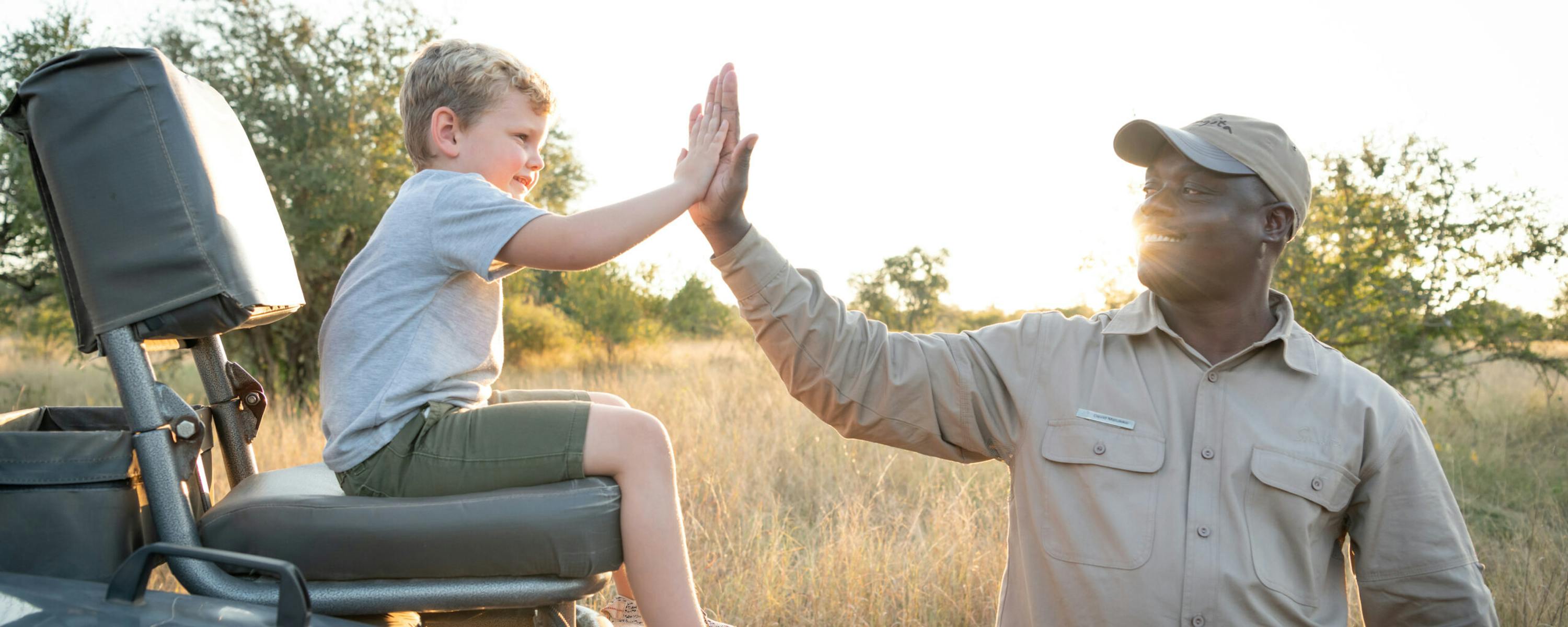 A safari guide high-fives a child seated in an open vehicle, with golden grassland and soft evening light behind.