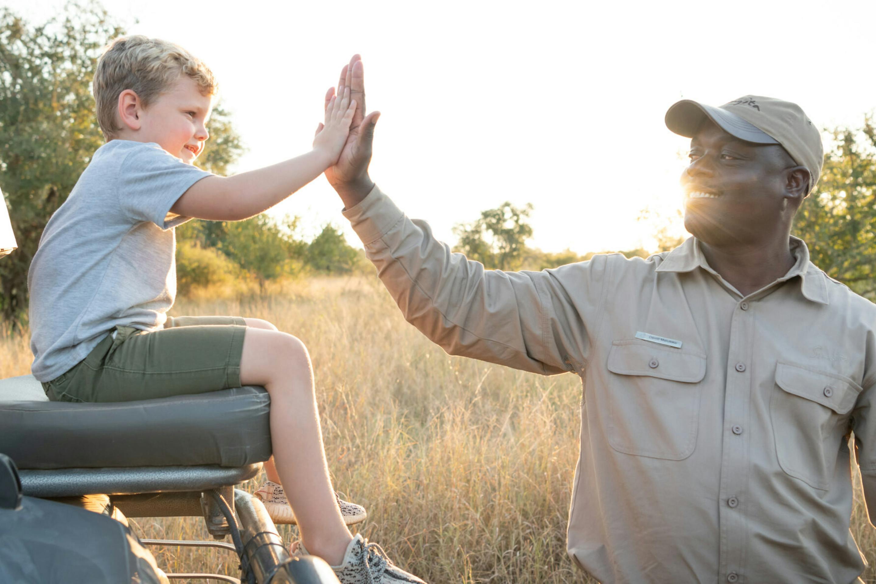 A safari guide high-fives a child seated in an open vehicle, with golden grassland and soft evening light behind.