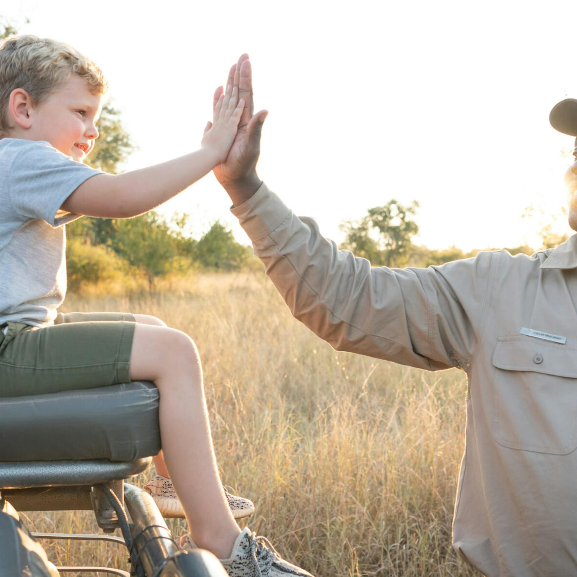 A safari guide high-fives a child seated in an open vehicle, with golden grassland and soft evening light behind.
